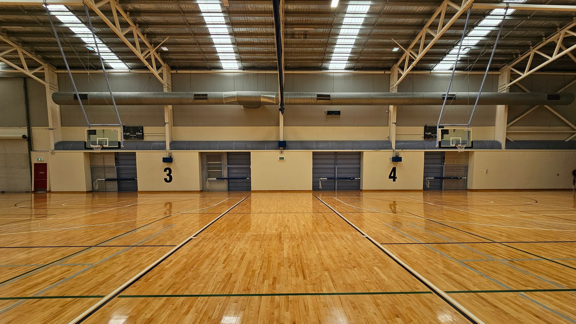 A spacious indoor basketball court with a polished wooden floor marked with lines. Two basketball hoops are visible on either side. The ceiling is high with metal beams and skylights. In the background, there is a kiosk area with tables and chairs, and a staircase leading to an upper level. The walls are a mix of brick and metal panelling. A spacious indoor basketball court with a polished wooden floor marked with lines. Two basketball hoops are visible on either side. The ceiling is high with metal beams and skylights. In the background, there is a kiosk area with tables and chairs, and a staircase leading to an upper level. The walls are a mix of brick and metal panelling.