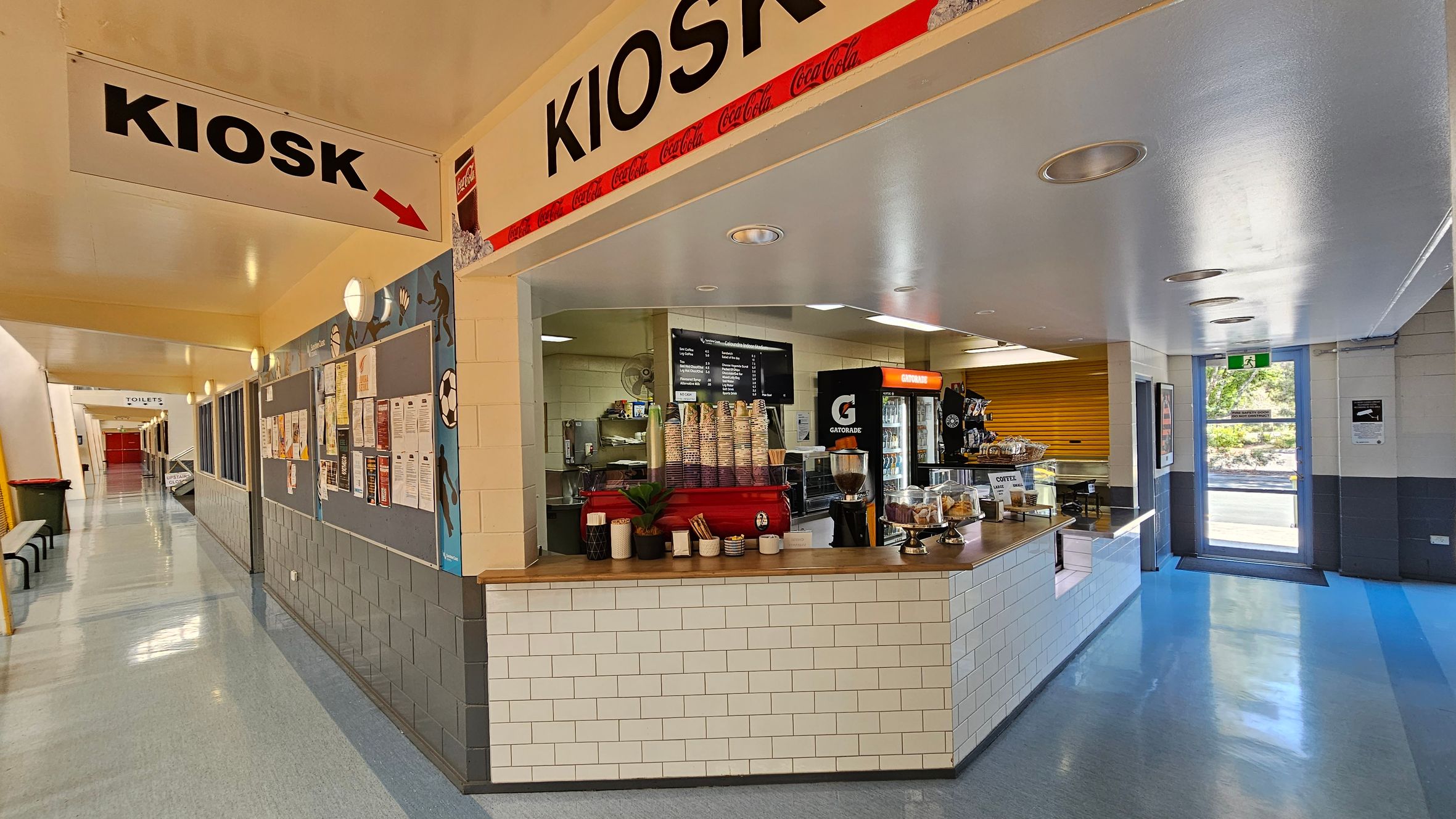 A sports hall with a polished wooden floor and a high ceiling with metal beams and skylights. In the background, there is a kiosk area with tables and chairs. A scoreboard is mounted above, displaying team scores. The wall features notice boards with various posters and information. A yellow pillar stands in the centre, and a staircase is visible on the right leading to an upper level. A green bin is on the left side. A sports hall with a polished wooden floor and a high ceiling with metal beams and skylights. In the background, there is a kiosk area with tables and chairs. A scoreboard is mounted above, displaying team scores. The wall features notice boards with various posters and information. A yellow pillar stands in the centre, and a staircase is visible on the right leading to an upper level. A green bin is on the left side.