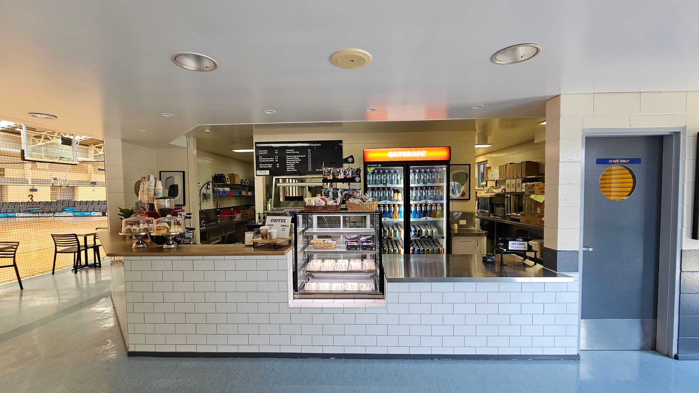 A café counter with a white tiled front. On the left, there are pastries and coffee cups on display. In the centre, a menu board is above a glass case with sandwiches. A fridge with a Gatorade sign is filled with drinks. To the right, a "Staff Only" door is visible. The background shows a sports court with seating and a net. The floor is a mix of grey and blue tiles, and the ceiling is white with recessed lighting. A café counter with a white tiled front. On the left, there are pastries and coffee cups on display. In the centre, a menu board is above a glass case with sandwiches. A fridge with a Gatorade sign is filled with drinks. To the right, a "Staff Only" door is visible. The background shows a sports court with seating and a net. The floor is a mix of grey and blue tiles, and the ceiling is white with recessed lighting.