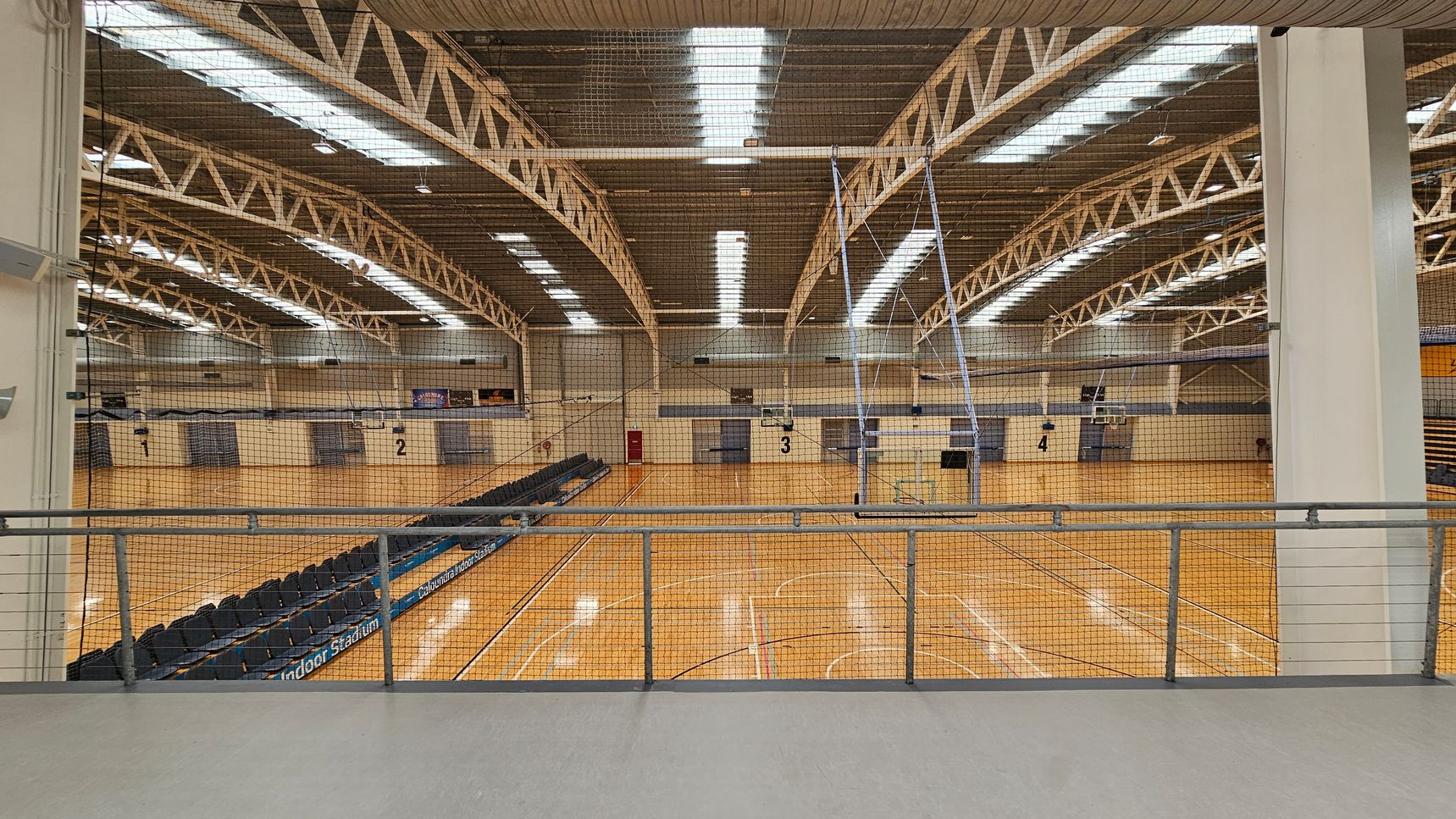 A large indoor sports hall with a wooden floor and multiple basketball courts marked with lines. The ceiling features exposed metal trusses and fluorescent lighting. On the left, there are rows of black seating. A netting barrier runs across the middle, separating the viewing area from the courts. The walls are light-coloured with numbered sections. A metal railing is in the foreground. A large indoor sports hall with a wooden floor and multiple basketball courts marked with lines. The ceiling features exposed metal trusses and fluorescent lighting. On the left, there are rows of black seating. A netting barrier runs across the middle, separating the viewing area from the courts. The walls are light-coloured with numbered sections. A metal railing is in the foreground.