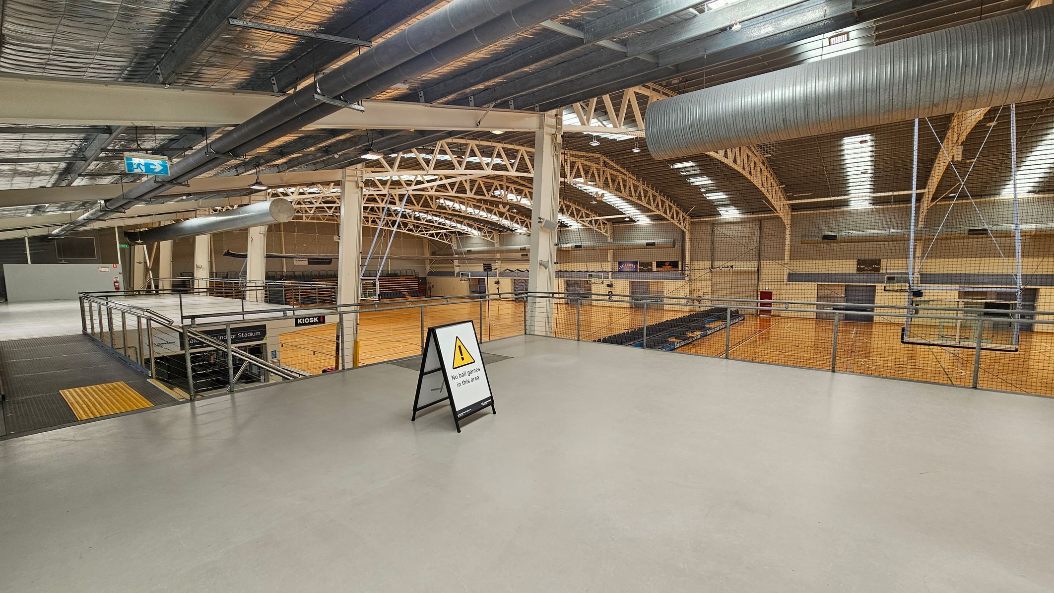 A large indoor sports hall with a high ceiling featuring exposed beams and ductwork. The floor is light grey, and a sign in the foreground reads "No ball games in this area." On the left, there is a staircase leading down, with a "Kiosk" sign visible. The background shows a wooden sports court with seating areas and a netted barrier. An exit sign is visible on the ceiling. A large indoor sports hall with a high ceiling featuring exposed beams and ductwork. The floor is light grey, and a sign in the foreground reads "No ball games in this area." On the left, there is a staircase leading down, with a "Kiosk" sign visible. The background shows a wooden sports court with seating areas and a netted barrier. An exit sign is visible on the ceiling.