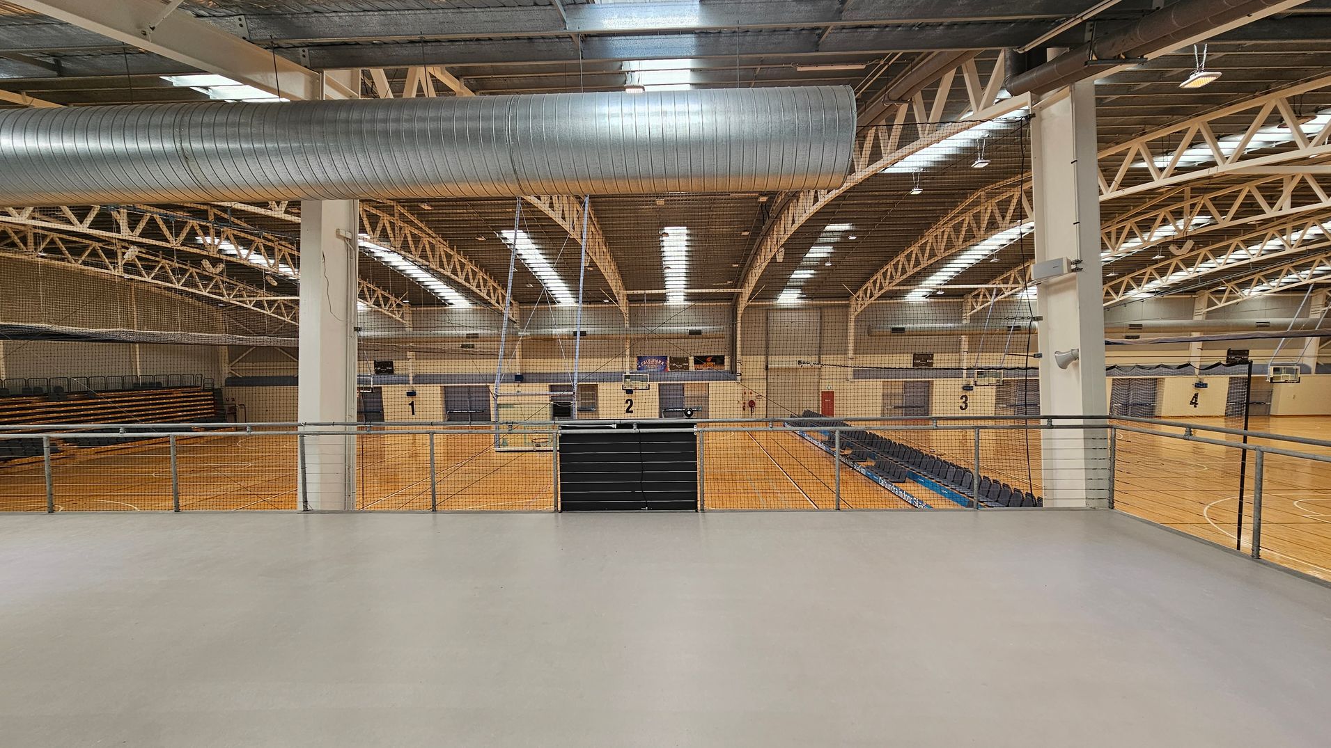 A large indoor sports hall with a high ceiling featuring exposed beams and a prominent metal ventilation duct. The floor is wooden, marked with lines for various sports. On the left, there are tiered seating areas, while on the right, more seating is visible. The hall is divided into multiple courts, numbered 2, 3, and 4, with basketball hoops and nets. A railing is in the foreground, separating a viewing area from the courts below. A large indoor sports hall with a high ceiling featuring exposed beams and a prominent metal ventilation duct. The floor is wooden, marked with lines for various sports. On the left, there are tiered seating areas, while on the right, more seating is visible. The hall is divided into multiple courts, numbered 2, 3, and 4, with basketball hoops and nets. A railing is in the foreground, separating a viewing area from the courts below.