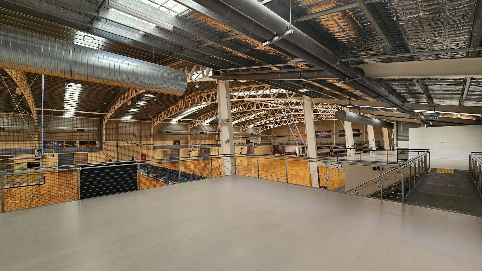 Interior of a large sports hall with a high ceiling featuring exposed metal beams and ductwork. The floor is wooden, marked for sports activities, and surrounded by seating areas. On the left, there are multiple courts with netting above. On the right, a mezzanine level with metal railings overlooks the courts. The ceiling has skylights allowing natural light in. A sign for toilets is visible on the right side near the stairs. Interior of a large sports hall with a high ceiling featuring exposed metal beams and ductwork. The floor is wooden, marked for sports activities, and surrounded by seating areas. On the left, there are multiple courts with netting above. On the right, a mezzanine level with metal railings overlooks the courts. The ceiling has skylights allowing natural light in. A sign for toilets is visible on the right side near the stairs.