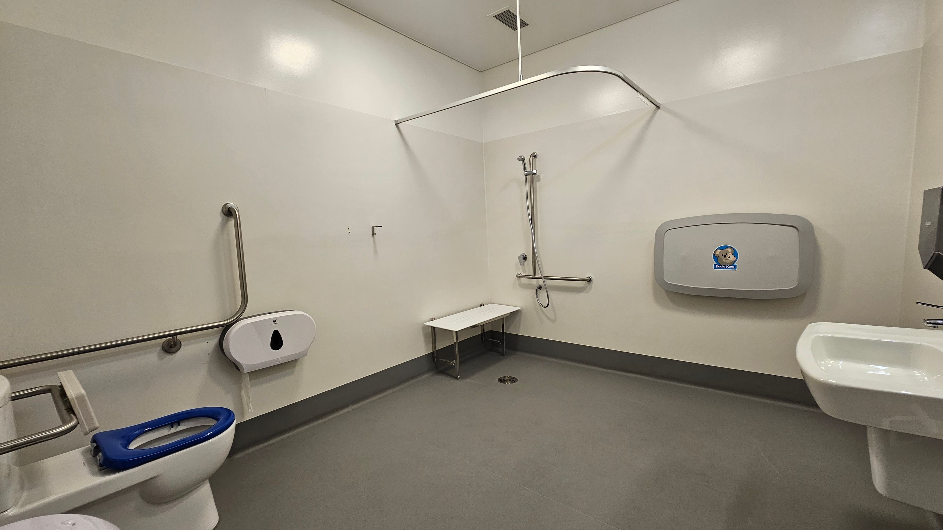 A spacious, accessible bathroom with a green floor and light-coloured walls. On the left, there is a toilet with a blue seat and a grab rail. In the centre, a shower area with a handheld showerhead and a foldable bench. On the right, a baby changing station is mounted on the wall above a white sink. The ceiling is plain with a light fixture. A spacious, accessible bathroom with a green floor and light-coloured walls. On the left, there is a toilet with a blue seat and a grab rail. In the centre, a shower area with a handheld showerhead and a foldable bench. On the right, a baby changing station is mounted on the wall above a white sink. The ceiling is plain with a light fixture.