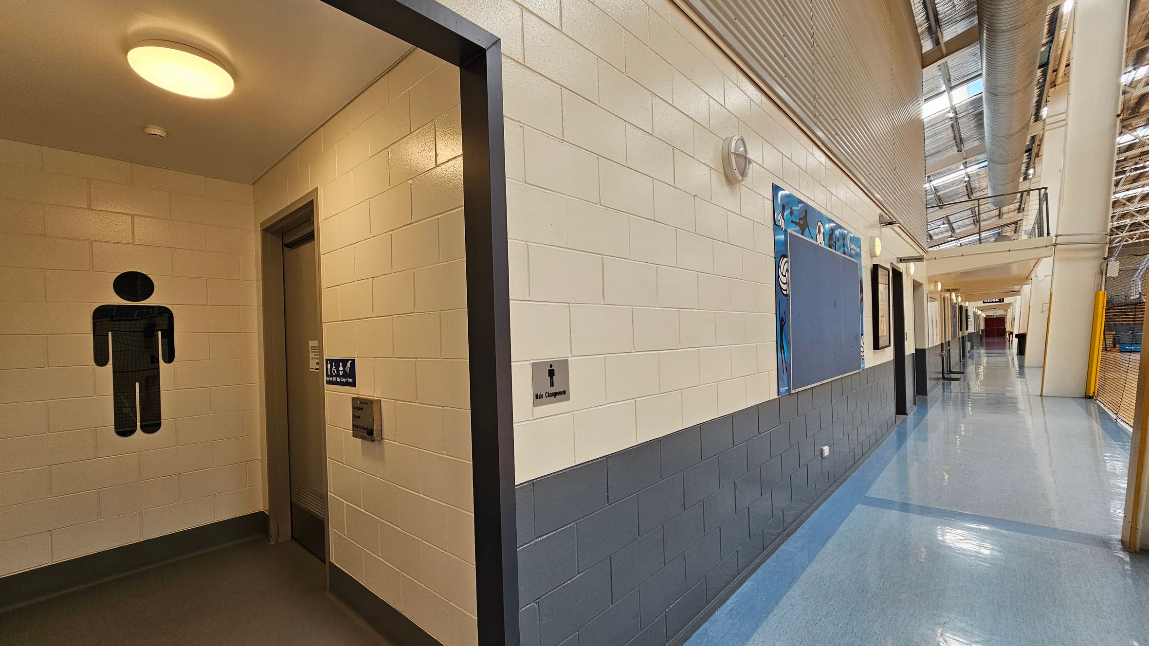 A corridor with a polished floor and a high ceiling. On the left, there is an entrance to a female changing room, marked by a symbol on the wall. Next to it is a closed roller shutter. The walls are tiled in a light colour with a darker base. On the right, a series of doors and a large noticeboard are visible. Ceiling lights illuminate the area, and a sign for "Kiosk 1" is seen further down the corridor. The ceiling has a metal structure with skylights. A corridor with a polished floor and a high ceiling. On the left, there is an entrance to a female changing room, marked by a symbol on the wall. Next to it is a closed roller shutter. The walls are tiled in a light colour with a darker base. On the right, a series of doors and a large noticeboard are visible. Ceiling lights illuminate the area, and a sign for "Kiosk 1" is seen further down the corridor. The ceiling has a metal structure with skylights.