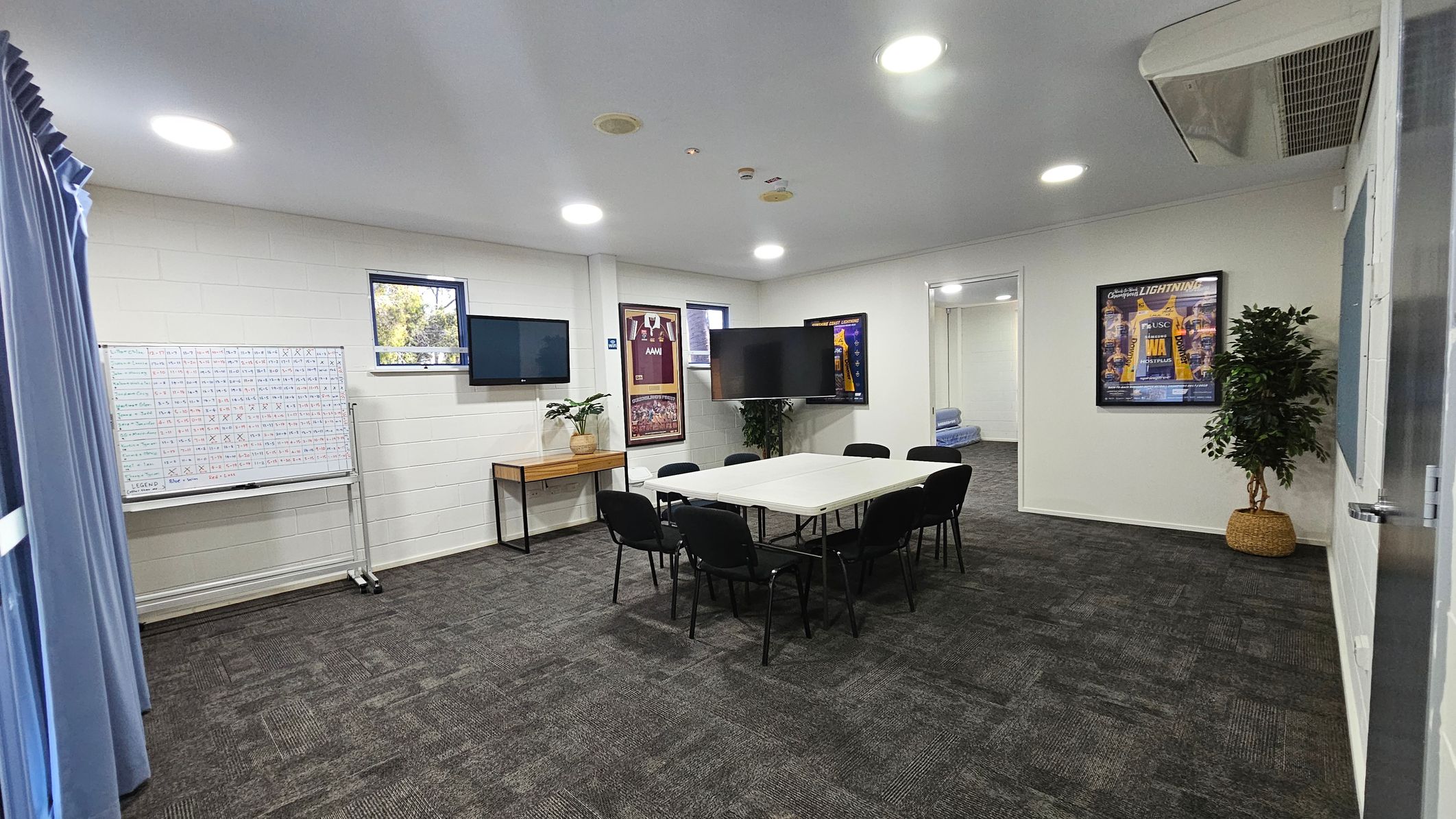 A hallway with a blue and grey tiled floor and cream and grey walls. On the left, an open door leads to a room with a whiteboard and a small window. A framed sports poster is on the wall next to the door. To the right, a closed grey roller shutter is visible. Further down the hallway, there are red double doors and a few black tables and chairs. The ceiling is high with visible beams. A hallway with a blue and grey tiled floor and cream and grey walls. On the left, an open door leads to a room with a whiteboard and a small window. A framed sports poster is on the wall next to the door. To the right, a closed grey roller shutter is visible. Further down the hallway, there are red double doors and a few black tables and chairs. The ceiling is high with visible beams.
