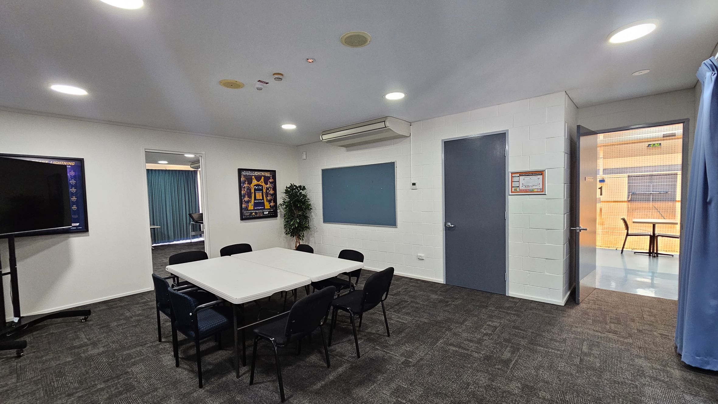 A meeting room with a rectangular table surrounded by black chairs on a dark carpeted floor. On the left, a large screen is mounted on a stand. A poster is on the wall next to a doorway leading to another room with blue curtains. In the centre, a blue notice board is mounted on the wall above a potted plant. On the right, a door is open, revealing a hallway with a table and chair. The ceiling has recessed lighting and an air conditioning unit. A meeting room with a rectangular table surrounded by black chairs on a dark carpeted floor. On the left, a large screen is mounted on a stand. A poster is on the wall next to a doorway leading to another room with blue curtains. In the centre, a blue notice board is mounted on the wall above a potted plant. On the right, a door is open, revealing a hallway with a table and chair. The ceiling has recessed lighting and an air conditioning unit.