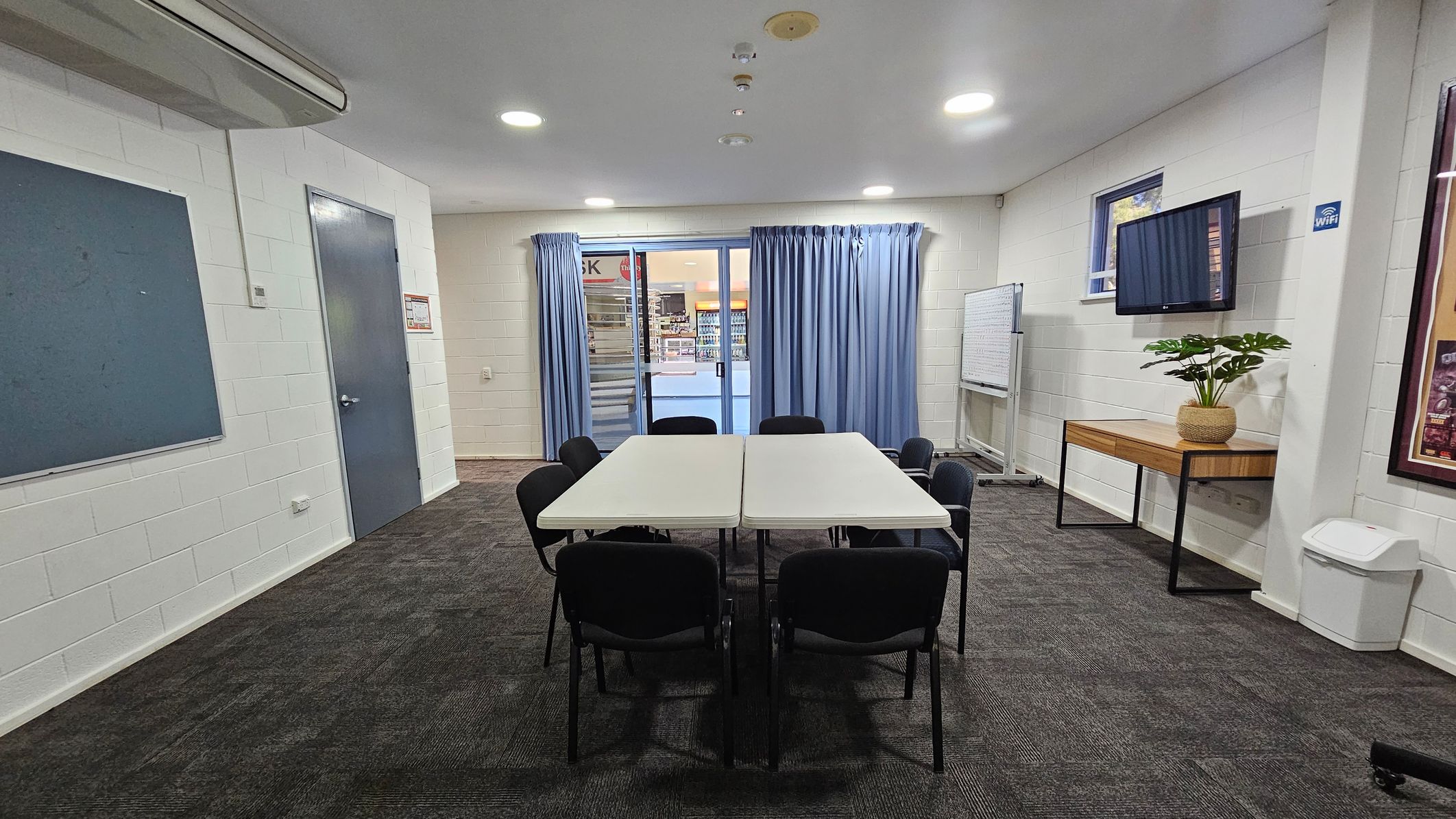 A room with a dark carpeted floor and white walls. In the centre, two rectangular tables are pushed together, surrounded by eight black chairs. On the right, a wooden table holds a potted plant beneath a wall-mounted TV. A whiteboard is nearby. The back wall features blue curtains covering a glass door, leading to a view of a shop. On the left, a grey door and a blue notice board are visible. The ceiling has recessed lighting and an air conditioning unit. A room with a dark carpeted floor and white walls. In the centre, two rectangular tables are pushed together, surrounded by eight black chairs. On the right, a wooden table holds a potted plant beneath a wall-mounted TV. A whiteboard is nearby. The back wall features blue curtains covering a glass door, leading to a view of a shop. On the left, a grey door and a blue notice board are visible. The ceiling has recessed lighting and an air conditioning unit.