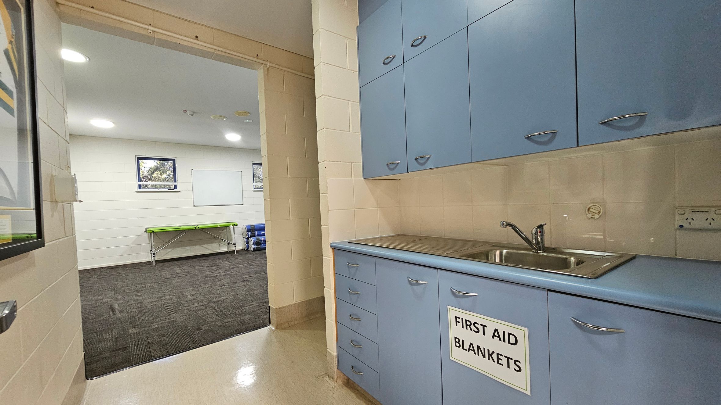 A room with light blue cabinets on the right, featuring a sink and a sign reading "First Aid Blankets." The floor is a light-coloured linoleum. Through an open doorway on the left, a room with dark carpet is visible, containing a green stretcher and rolled-up mats. The walls are cream-coloured with two small windows and a whiteboard. The ceiling has recessed lighting. A room with light blue cabinets on the right, featuring a sink and a sign reading "First Aid Blankets." The floor is a light-coloured linoleum. Through an open doorway on the left, a room with dark carpet is visible, containing a green stretcher and rolled-up mats. The walls are cream-coloured with two small windows and a whiteboard. The ceiling has recessed lighting.