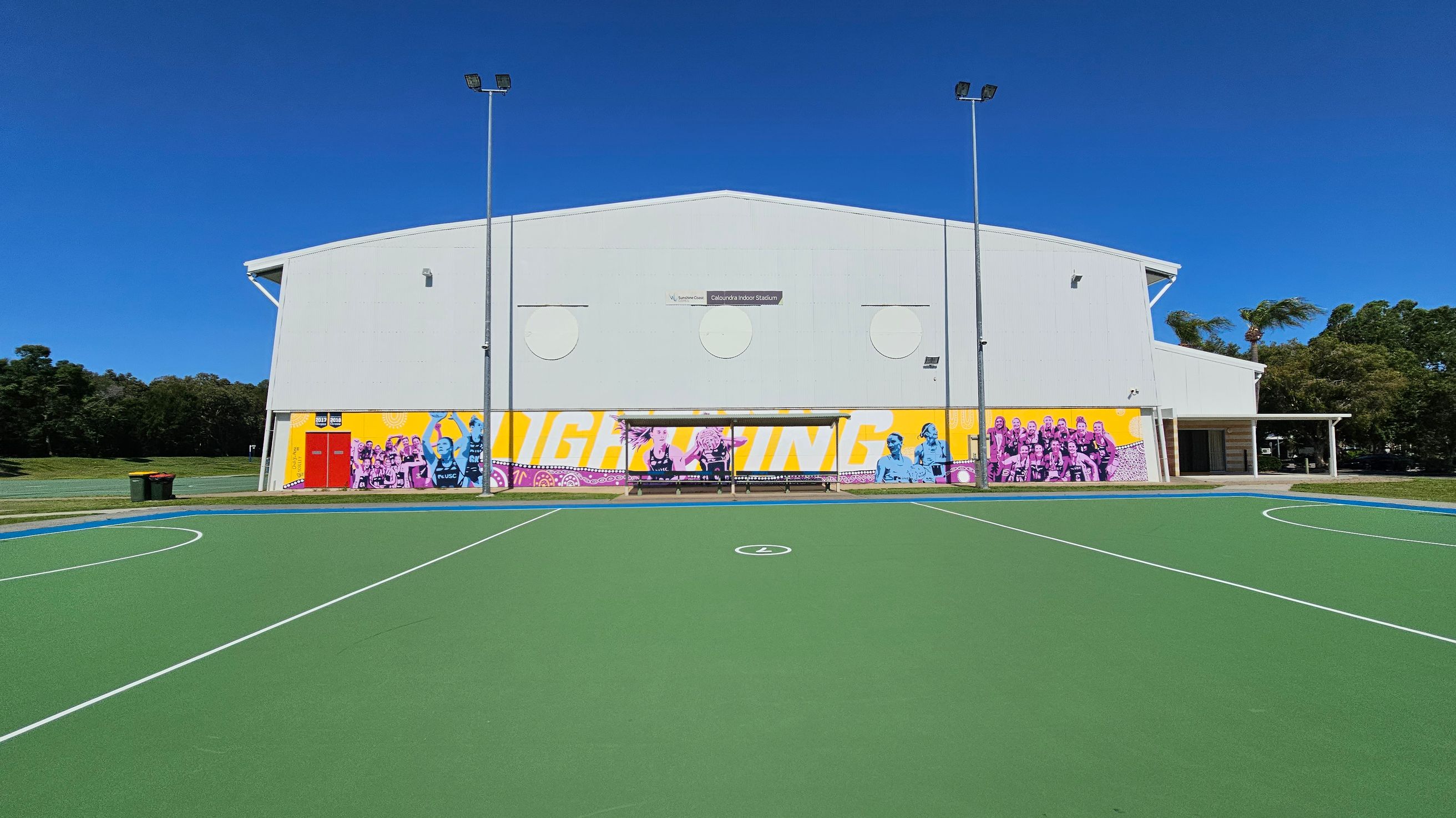 A sports court with a green surface and white lines in the foreground. In the background, a large white building with colourful murals featuring people and text. The building has a sign reading "Caloundra Indoor Stadium" and is flanked by tall light poles. To the left, there are green bins and grass, while trees are visible on both sides. A sports court with a green surface and white lines in the foreground. In the background, a large white building with colourful murals featuring people and text. The building has a sign reading "Caloundra Indoor Stadium" and is flanked by tall light poles. To the left, there are green bins and grass, while trees are visible on both sides.