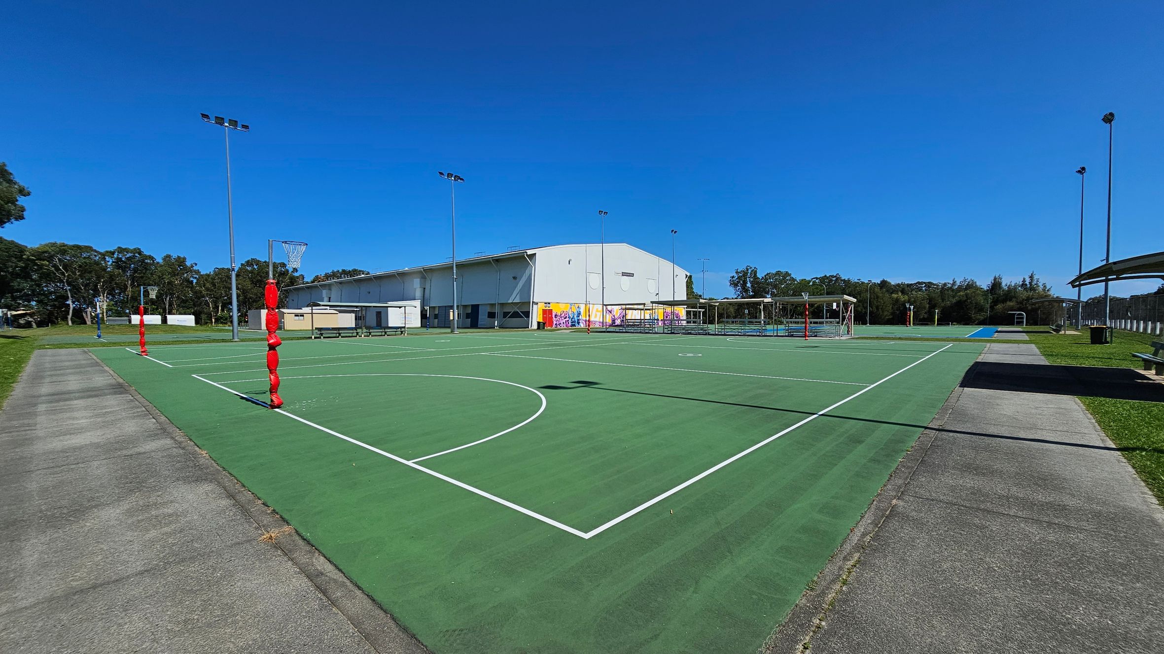 A green outdoor netball court with white lines is in the centre. Red padding covers the goal posts. On the left, a concrete path runs alongside the court, bordered by grass. In the background, a large building with colourful murals is visible. Several tall floodlights surround the area. On the right, there are more courts and a covered seating area. Trees line the horizon. A green outdoor netball court with white lines is in the centre. Red padding covers the goal posts. On the left, a concrete path runs alongside the court, bordered by grass. In the background, a large building with colourful murals is visible. Several tall floodlights surround the area. On the right, there are more courts and a covered seating area. Trees line the horizon.