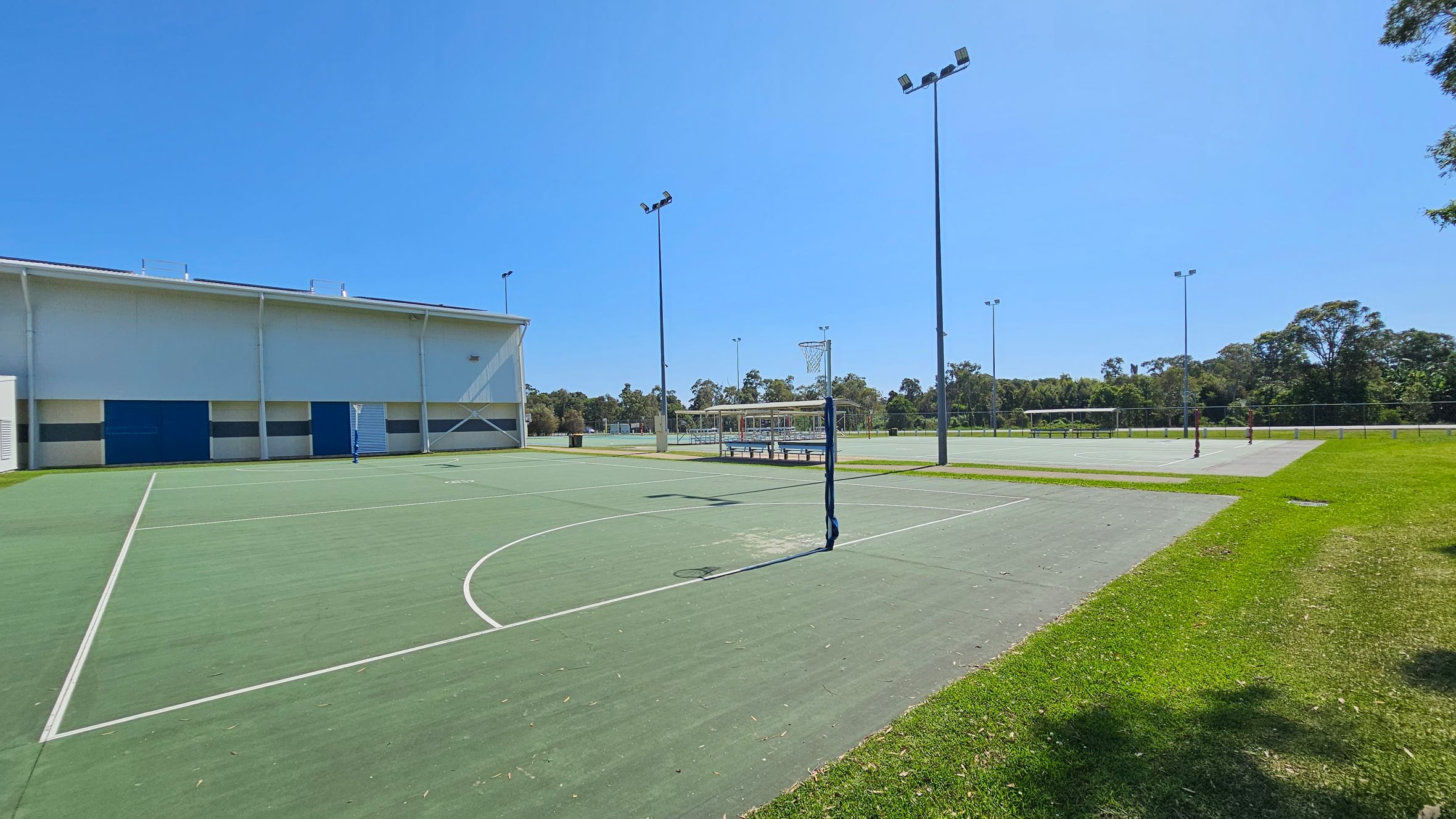 Outdoor sports courts with green surfaces and white lines. On the left, a large building with a white and blue exterior. In the centre, a netball hoop and tall floodlights. To the right, more courts and a grassy area. Trees and a fence line the background. Outdoor sports courts with green surfaces and white lines. On the left, a large building with a white and blue exterior. In the centre, a netball hoop and tall floodlights. To the right, more courts and a grassy area. Trees and a fence line the background.