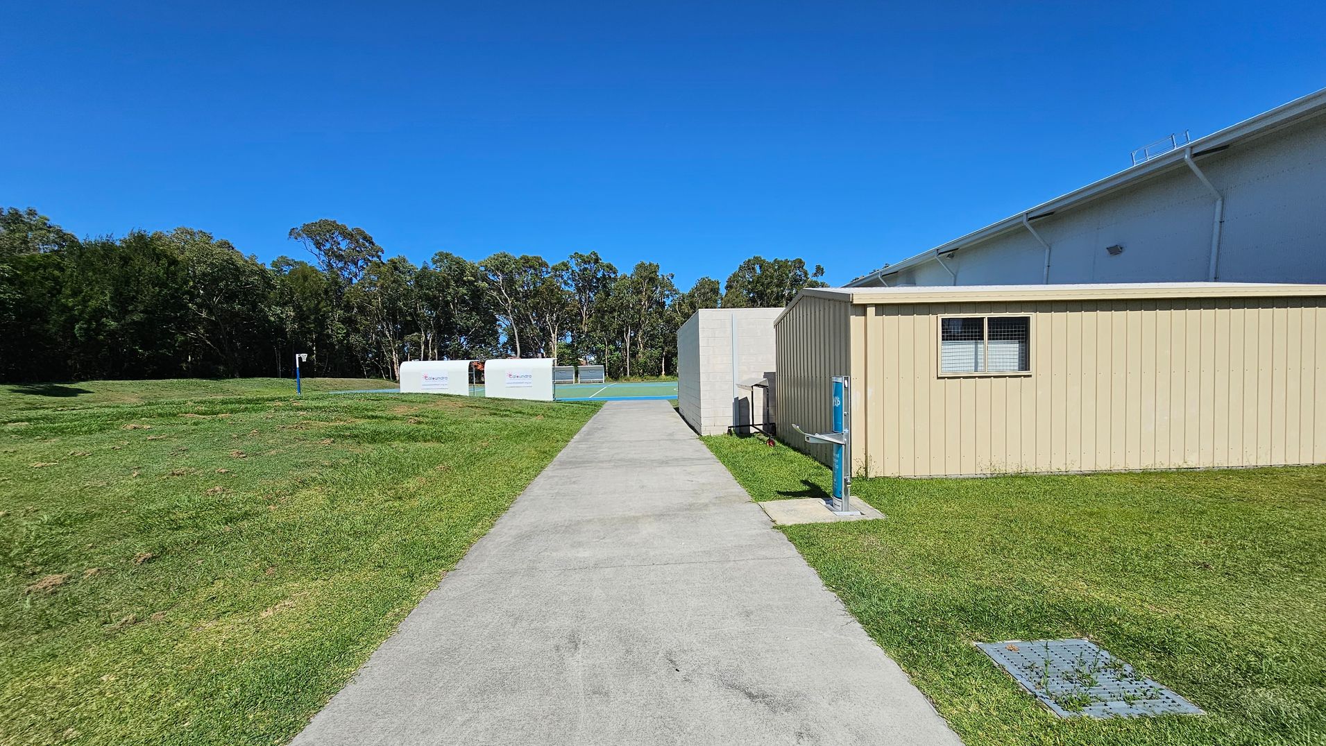 A concrete path runs through the centre, flanked by green grass. On the right, there's a beige building with a small window. To the left, open grassy fields extend towards a line of trees in the background. In the distance, there are two white structures with signage. The sky is clear and blue. A concrete path runs through the centre, flanked by green grass. On the right, there's a beige building with a small window. To the left, open grassy fields extend towards a line of trees in the background. In the distance, there are two white structures with signage. The sky is clear and blue.