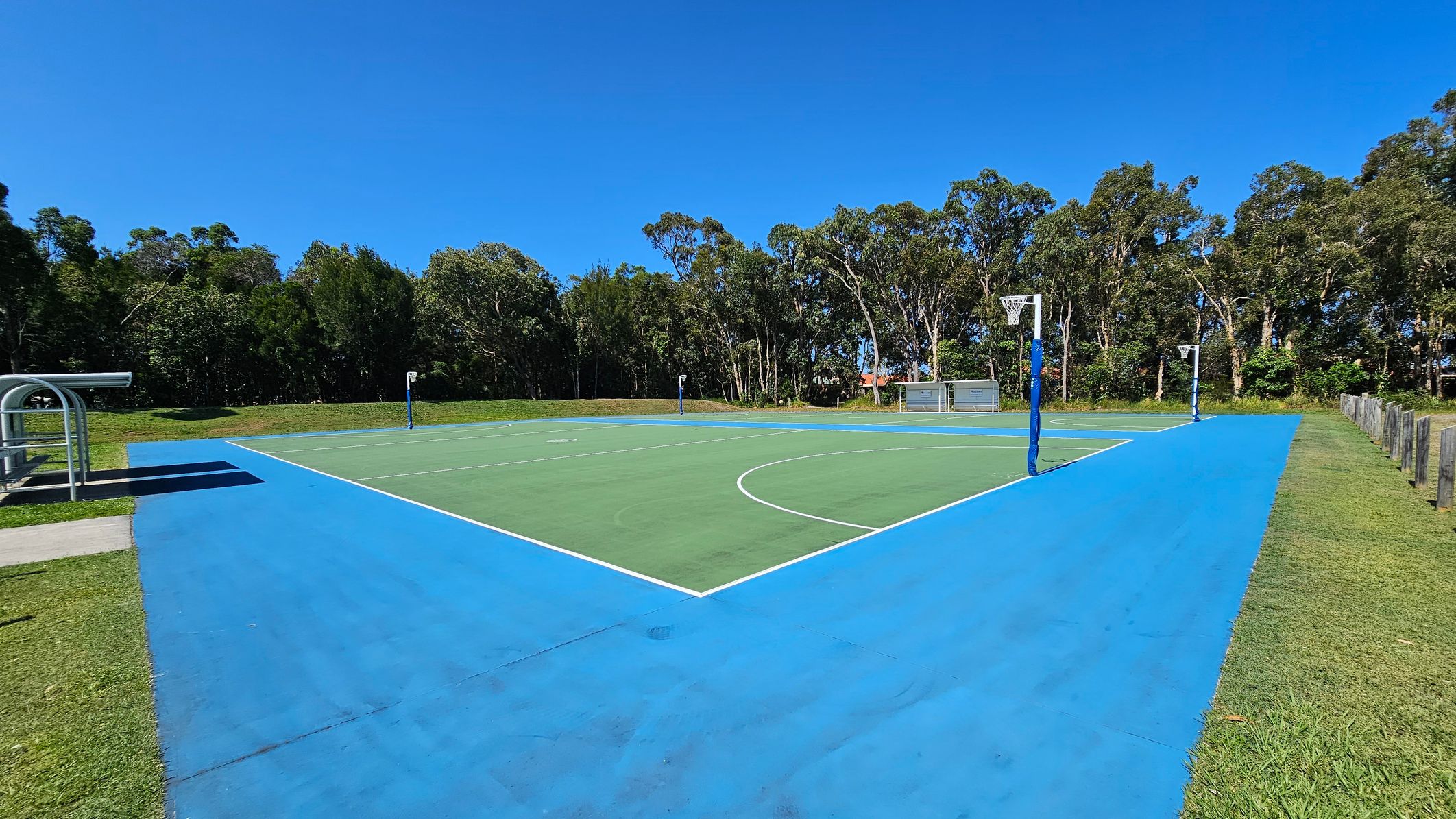 A netball court with a green playing area and a wide blue border. On the left, there's a covered bench area with a metal frame. The right side has a grassy edge with wooden posts. In the background, a line of tall trees provides a natural backdrop. The court has two netball hoops, one on each end. A netball court with a green playing area and a wide blue border. On the left, there's a covered bench area with a metal frame. The right side has a grassy edge with wooden posts. In the background, a line of tall trees provides a natural backdrop. The court has two netball hoops, one on each end.
