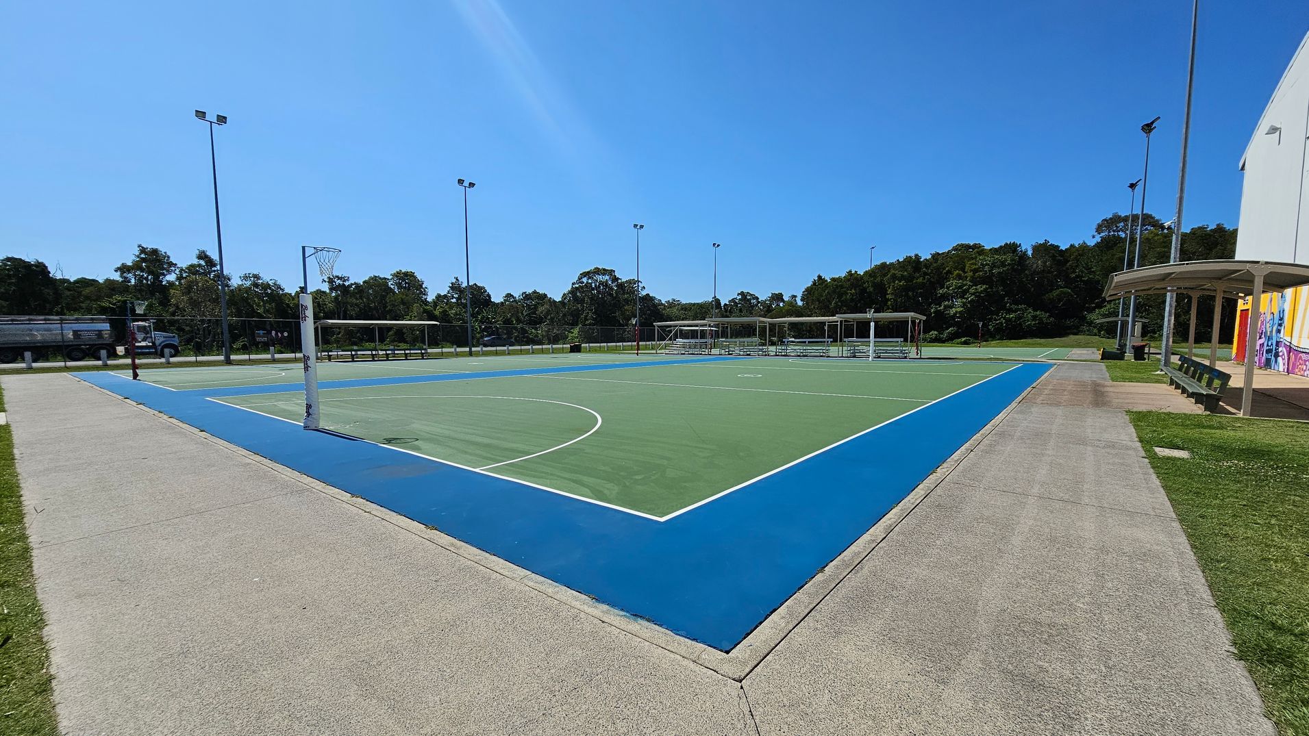 A netball court with a green playing surface and a blue border. On the left, a concrete path runs alongside the court, with a truck visible in the background near some trees. On the right, there is a covered seating area with benches and a building with colourful murals. Tall floodlights surround the court, and trees line the background. A netball court with a green playing surface and a blue border. On the left, a concrete path runs alongside the court, with a truck visible in the background near some trees. On the right, there is a covered seating area with benches and a building with colourful murals. Tall floodlights surround the court, and trees line the background.