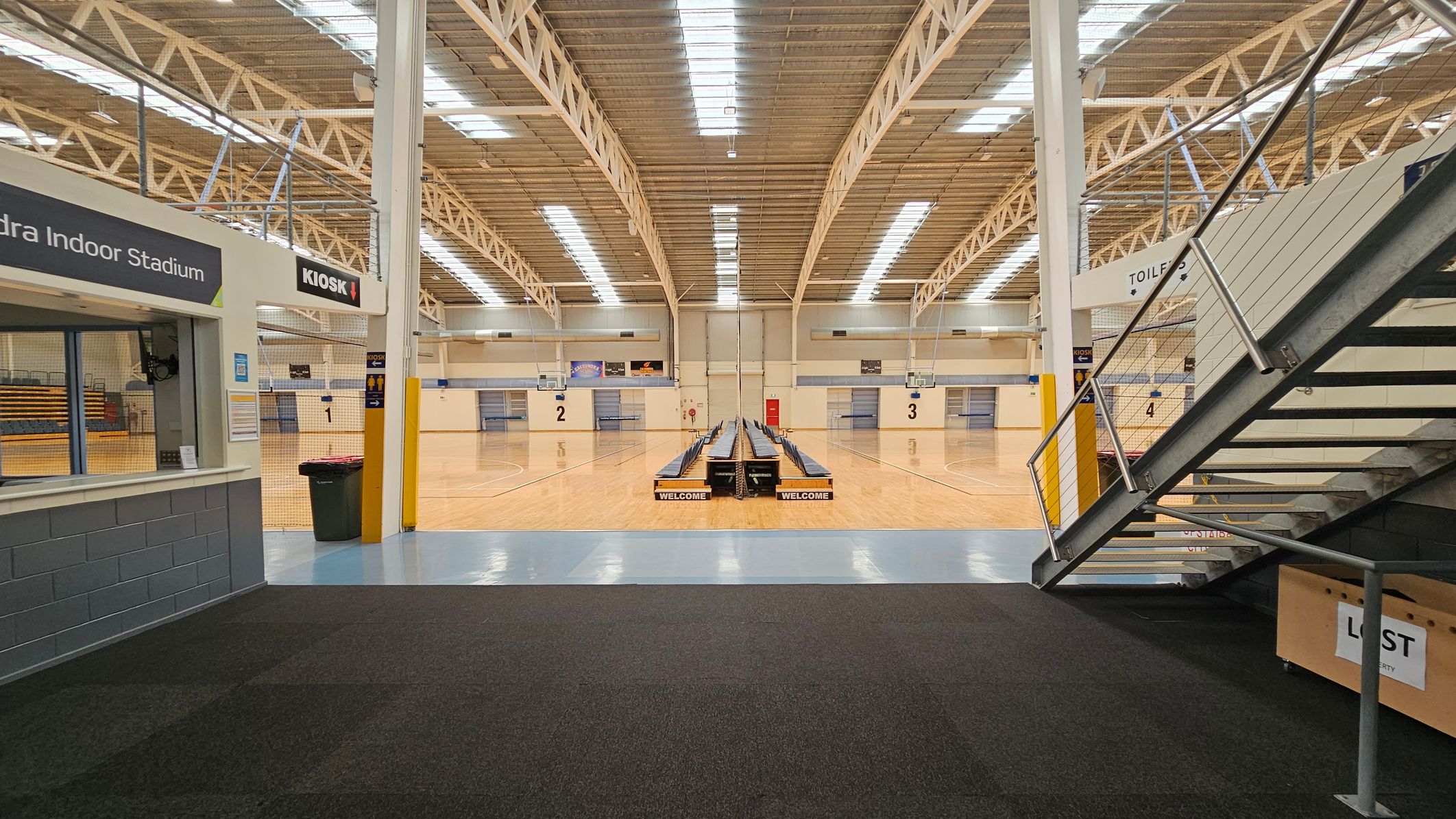 Glass entrance with blue framing, leading into a building. On the left, a yellow poster with social media information. On the right, a sign with entry conditions. Inside, a reception desk is visible, with a staircase to the right. The floor is tiled, and the ceiling has recessed lighting. Outside, a concrete path and some greenery are visible. Glass entrance with blue framing, leading into a building. On the left, a yellow poster with social media information. On the right, a sign with entry conditions. Inside, a reception desk is visible, with a staircase to the right. The floor is tiled, and the ceiling has recessed lighting. Outside, a concrete path and some greenery are visible.