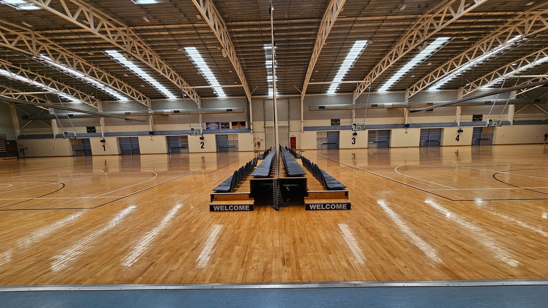 A large indoor sports hall with a polished wooden floor. In the centre, there are two rows of blue chairs facing opposite directions, with "WELCOME" signs at the front. The ceiling features exposed metal beams and skylights. The walls are beige with numbered sections from 1 to 4, each with a basketball hoop. The space is well-lit, and the floor markings indicate multiple basketball courts. A large indoor sports hall with a polished wooden floor. In the centre, there are two rows of blue chairs facing opposite directions, with "WELCOME" signs at the front. The ceiling features exposed metal beams and skylights. The walls are beige with numbered sections from 1 to 4, each with a basketball hoop. The space is well-lit, and the floor markings indicate multiple basketball courts.