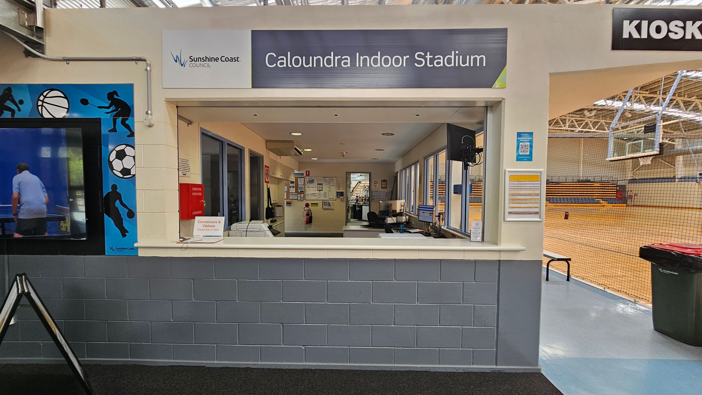 Reception area of Caloundra Indoor Stadium. The counter has a sign for contractors and visitors. Behind the counter are office equipment and notice boards. To the left, a wall features sports-themed graphics. On the right, an entrance leads to a gymnasium with a basketball hoop and bleachers. The ceiling has exposed beams and lights, and the floor is a mix of carpet and polished surface. A kiosk sign is visible above the entrance. Reception area of Caloundra Indoor Stadium. The counter has a sign for contractors and visitors. Behind the counter are office equipment and notice boards. To the left, a wall features sports-themed graphics. On the right, an entrance leads to a gymnasium with a basketball hoop and bleachers. The ceiling has exposed beams and lights, and the floor is a mix of carpet and polished surface. A kiosk sign is visible above the entrance.