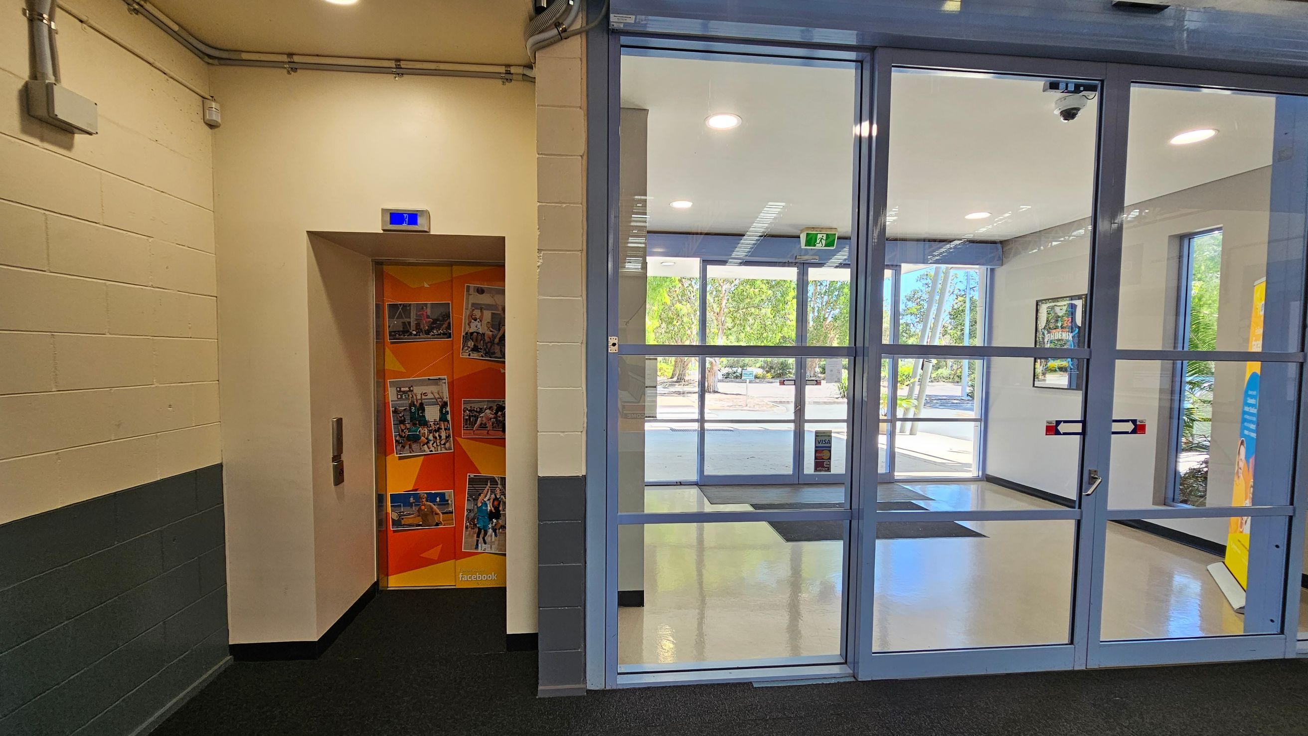 On the left, there's a lift with an orange door featuring various photos. The wall is cream with a grey lower section, and there are visible pipes near the ceiling. On the right, a glass entrance door leads to a bright lobby with large windows showing trees outside. The floor is a mix of dark carpet and light tiles. A poster is partially visible on the far right. On the left, there's a lift with an orange door featuring various photos. The wall is cream with a grey lower section, and there are visible pipes near the ceiling. On the right, a glass entrance door leads to a bright lobby with large windows showing trees outside. The floor is a mix of dark carpet and light tiles. A poster is partially visible on the far right.