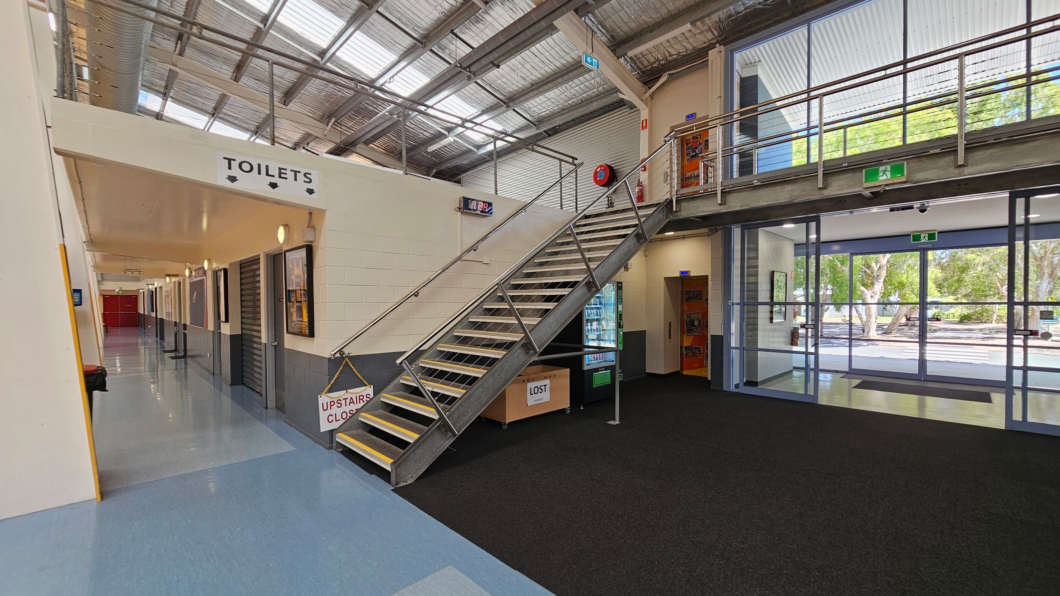 A modern indoor space with a metal staircase in the centre leading to a mezzanine level. To the left, a corridor with a sign for toilets and several doors. The floor is a mix of blue and black tiles. On the right, large glass doors and windows provide a view of trees outside. A vending machine and a "Lost" box are near the staircase. The ceiling is industrial with exposed metal beams and skylights. A modern indoor space with a metal staircase in the centre leading to a mezzanine level. To the left, a corridor with a sign for toilets and several doors. The floor is a mix of blue and black tiles. On the right, large glass doors and windows provide a view of trees outside. A vending machine and a "Lost" box are near the staircase. The ceiling is industrial with exposed metal beams and skylights.