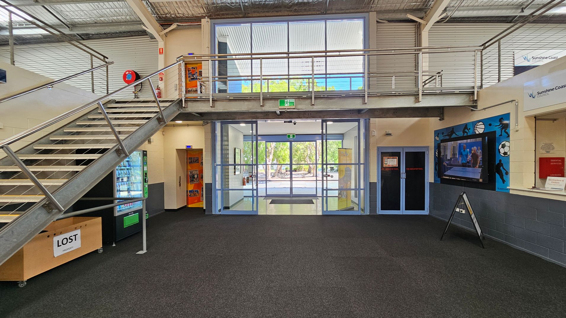 A spacious indoor area with a dark carpeted floor. On the left, a metal staircase leads to an upper level with a railing. Below the stairs, there's a vending machine and a "Lost Property" box. In the centre, large glass doors open to an outdoor area with trees visible. On the right, a wall-mounted TV displays a sports scene, and there's a sign for "Sunshine Coast Council" above. The ceiling is industrial with exposed beams and ductwork. A spacious indoor area with a dark carpeted floor. On the left, a metal staircase leads to an upper level with a railing. Below the stairs, there's a vending machine and a "Lost Property" box. In the centre, large glass doors open to an outdoor area with trees visible. On the right, a wall-mounted TV displays a sports scene, and there's a sign for "Sunshine Coast Council" above. The ceiling is industrial with exposed beams and ductwork.