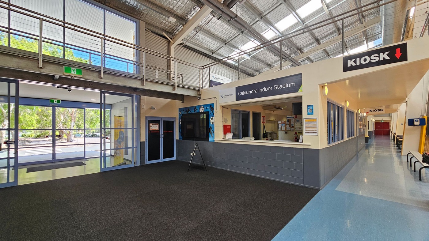 Interior of a building with a high ceiling featuring exposed metal beams and skylights. On the left, large glass doors lead outside, showing trees through the windows. In the centre, a reception area for "Caloundra Indoor Stadium" with a counter and signage. To the right, a corridor with a "Kiosk" sign above, leading to a red door. The floor is a mix of dark carpet and light blue tiles, and there are benches along the corridor. Interior of a building with a high ceiling featuring exposed metal beams and skylights. On the left, large glass doors lead outside, showing trees through the windows. In the centre, a reception area for "Caloundra Indoor Stadium" with a counter and signage. To the right, a corridor with a "Kiosk" sign above, leading to a red door. The floor is a mix of dark carpet and light blue tiles, and there are benches along the corridor.