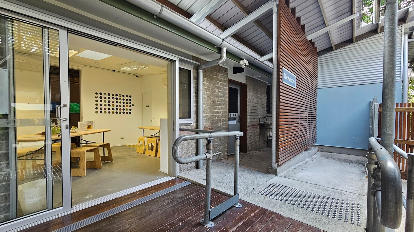 A modern building entrance with a sliding glass door on the left, revealing a room with wooden tables and stools. The floor inside is light grey, and the walls are white with a circular pattern decoration. Outside, there's a wooden deck and a metal railing. On the right, a brick wall and a wooden slatted panel with a sign reading "The Anthoni" are visible. The ceiling is metal with exposed beams, and the exterior wall is partly blue.