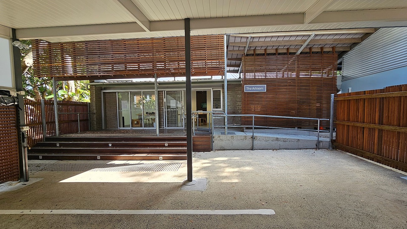 A covered outdoor area with a textured concrete floor and a white ceiling. In the centre, a metal pole supports the structure. Wooden slats form a partial wall on the left and right, with a wooden fence extending on both sides. In the background, a building with sliding glass doors and a sign reading "The Artroom" is visible. Steps lead up to the entrance, and a metal railing runs along the right side. Trees and foliage can be seen through the slats on the left.