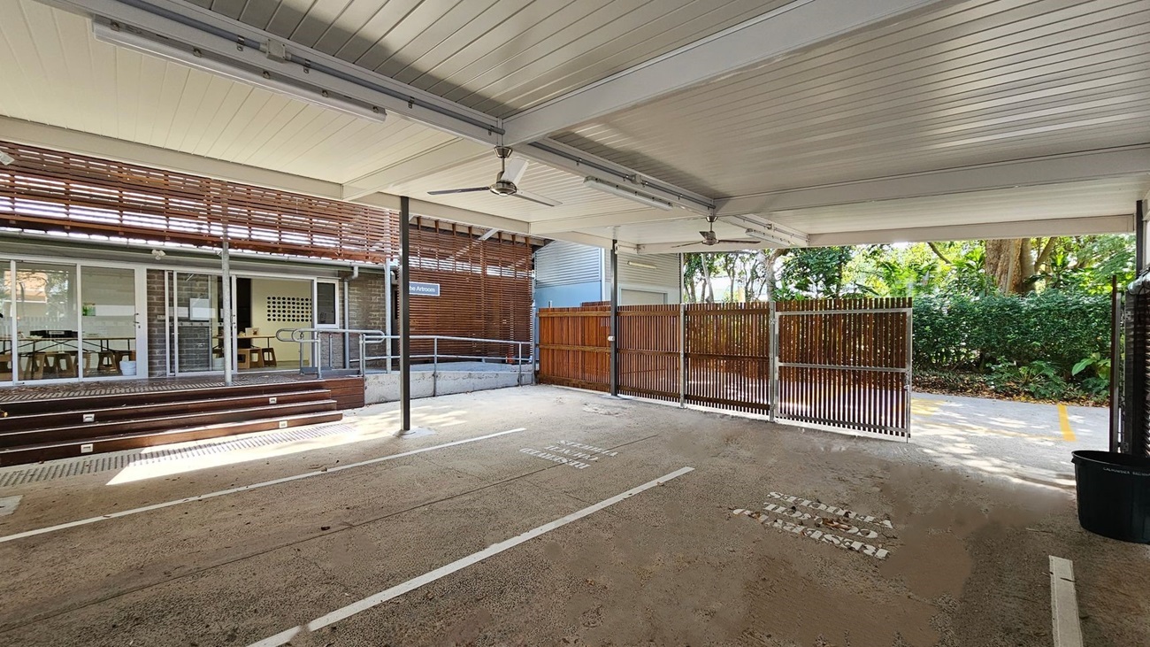 Covered outdoor area with a concrete floor, scattered with dry leaves. The ceiling is lined with panels and has ceiling fans. On the left, there are glass doors and windows leading to an indoor space with tables and chairs. A wooden ramp and steps provide access. On the right, a wooden fence and greenery are visible. The background shows trees and bushes, creating a natural setting.