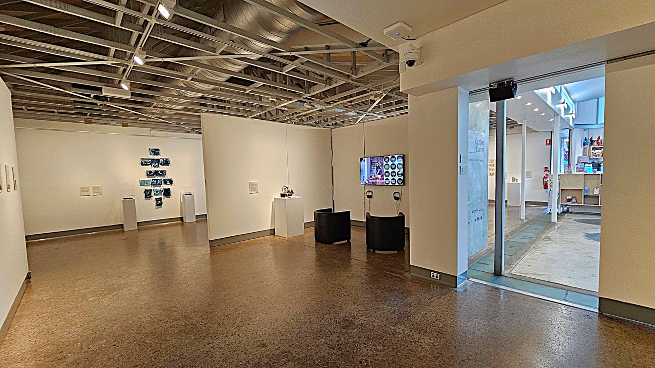An art gallery interior with white walls and a polished brown floor. On the left, a series of blue artworks are displayed on the wall. In the centre, a small sculpture is placed on a pedestal. Two black chairs face a wall-mounted screen with headphones. The ceiling features exposed ductwork and lighting. On the right, a glass door leads to another room with shelves and a fire extinguisher visible.