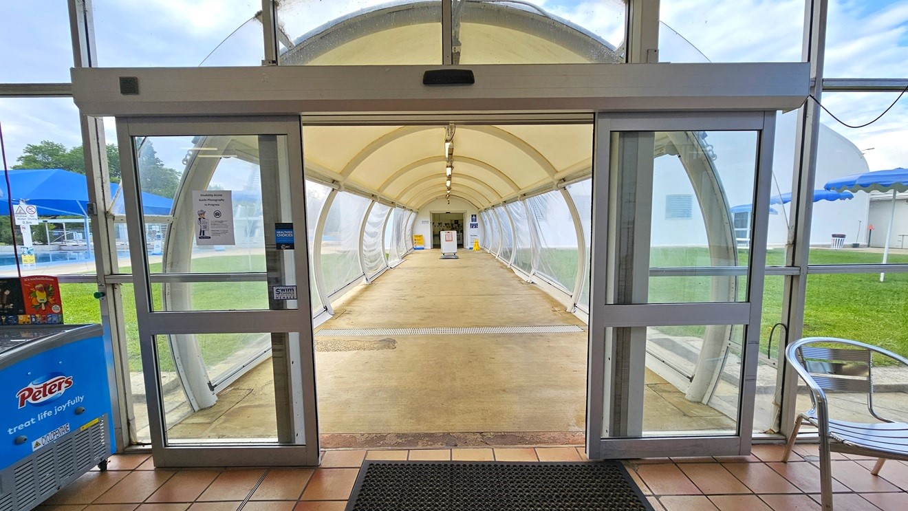 In the middle is an automatic glass door that leads to a tunnel constructed with durable vinyl material, featuring adjustable side panel flaps. An anti-slip mat is placed on the doorsteps and movable silver chairs are visible on the right. On the left is an ice cream freezer. Both sides of the tunnel are green grass and blue shade cloth. Truncated domes are installed near the door. Further ahead, at the end of the tunnel, banners are visible. In the middle is an automatic glass door that leads to a tunnel constructed with durable vinyl material, featuring adjustable side panel flaps. An anti-slip mat is placed on the doorsteps and movable silver chairs are visible on the right. On the left is an ice cream freezer. Both sides of the tunnel are green grass and blue shade cloth. Truncated domes are installed near the door. Further ahead, at the end of the tunnel, banners are visible.