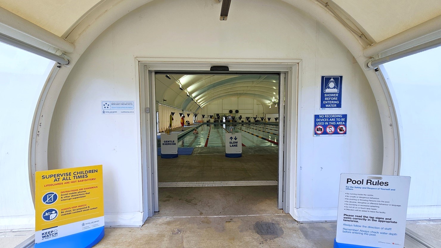 The dome-shaped entrance displays posters and safety reminders. Inside, truncated domes are installed. The lap pool is visible and equipped with floating ropes. Free-standing banners are positioned on the pool deck. The dome-shaped entrance displays posters and safety reminders. Inside, truncated domes are installed. The lap pool is visible and equipped with floating ropes. Free-standing banners are positioned on the pool deck.