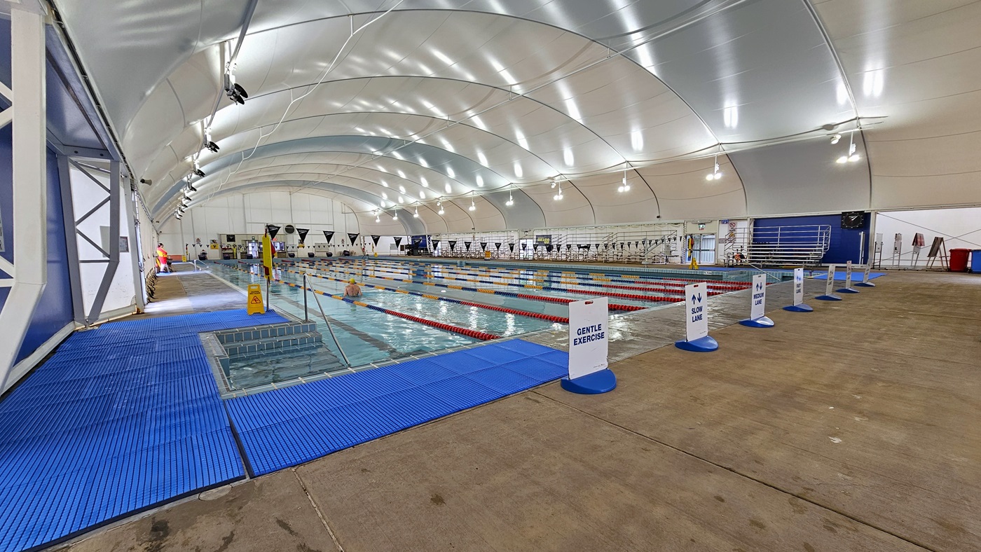 In the middle is the 8-lane lap pool, equipped with floating ropes and stairs with railings. Free-standing banners are positioned on the pool deck. Blue matting is installed in the left corner. Tiered seatings are positioned on the right. Strings of blue flags are hung across the pool. The dome-shaped ceiling features floodlights and LED lights In the middle is the 8-lane lap pool, equipped with floating ropes and stairs with railings. Free-standing banners are positioned on the pool deck. Blue matting is installed in the left corner. Tiered seatings are positioned on the right. Strings of blue flags are hung across the pool. The dome-shaped ceiling features floodlights and LED lights