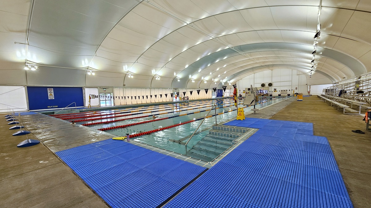 In the center is the 8-lane lap pool, equipped with floating ropes and stairs with railings. Free-standing banners are positioned on the pool deck. Blue matting is installed on both corners. Tiered seating is on the right. Strings of blue flags are hung across the pool. Safety reminder banners are visible on the pool deck area to the right. The dome-shaped ceiling features floodlights and LED lights. In the center is the 8-lane lap pool, equipped with floating ropes and stairs with railings. Free-standing banners are positioned on the pool deck. Blue matting is installed on both corners. Tiered seating is on the right. Strings of blue flags are hung across the pool. Safety reminder banners are visible on the pool deck area to the right. The dome-shaped ceiling features floodlights and LED lights.