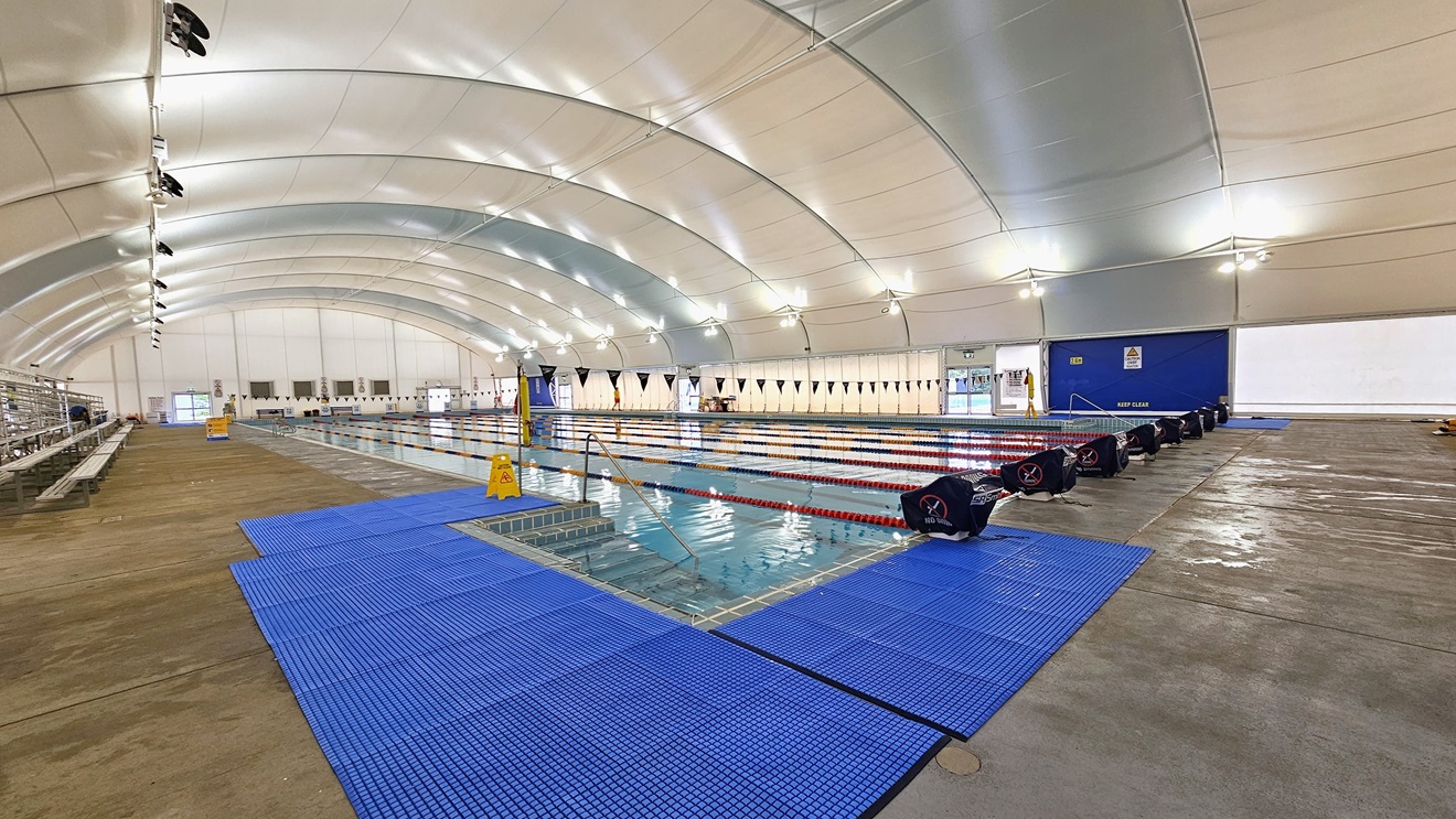 In the center is the 8-lane lap pool, equipped with floating ropes and stairs with railings. Diving boards are arranged on the pool deck. Blue matting is installed on both corners. Tiered seating is on the right. Strings of blue flags are hung across the pool. Tiered seating and safety reminder banners are visible on the pool deck area to the left. The dome-shaped ceiling features floodlights and LED lights. In the center is the 8-lane lap pool, equipped with floating ropes and stairs with railings. Diving boards are arranged on the pool deck. Blue matting is installed on both corners. Tiered seating is on the right. Strings of blue flags are hung across the pool. Tiered seating and safety reminder banners are visible on the pool deck area to the left. The dome-shaped ceiling features floodlights and LED lights.