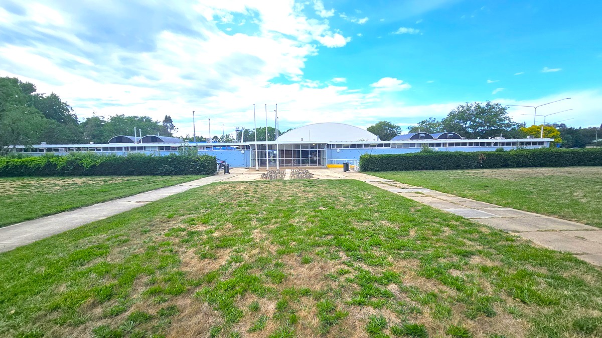 In the centre stands a concrete building with a dome-shaped roof. Trimmed shrubs border the front of the building. Adjacent to it lies expansive ticketed grassy parkland. Two concrete pathways lead to the building's entrance, and a modern concrete bike rack is positioned towards the end of the car park. Multiple street lights and trees surround the area. In the centre stands a concrete building with a dome-shaped roof. Trimmed shrubs border the front of the building. Adjacent to it lies expansive ticketed grassy parkland. Two concrete pathways lead to the building's entrance, and a modern concrete bike rack is positioned towards the end of the car park. Multiple street lights and trees surround the area.