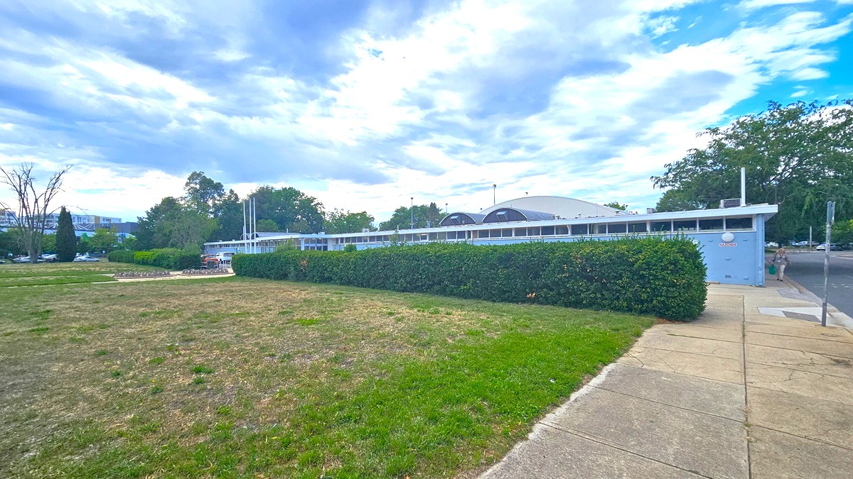 To the left, an expansive grassy parkland extends. On the right, a brick pathway and a road with a step-free entrance are present. Behind stands a blue concrete building with a dome-shaped roof. Trimmed ornamental shrubs border the building's front. Towards the far left, a concrete bike rack is positioned. The area is surrounded by trees. To the left, an expansive grassy parkland extends. On the right, a brick pathway and a road with a step-free entrance are present. Behind stands a blue concrete building with a dome-shaped roof. Trimmed ornamental shrubs border the building's front. Towards the far left, a concrete bike rack is positioned. The area is surrounded by trees.
