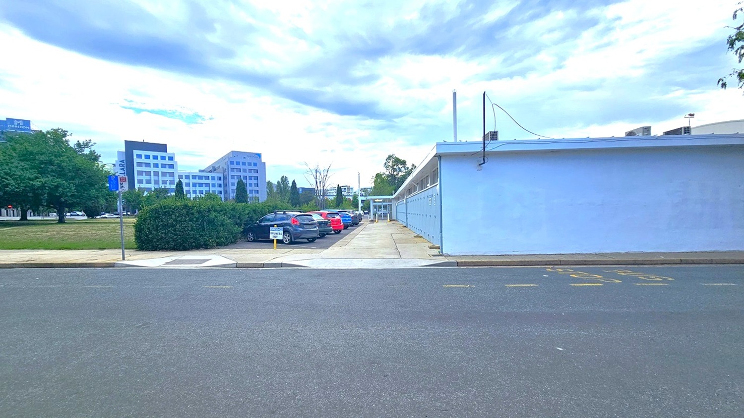 To the left lies an expansive grassy parkland, while on the right, a brick pathway and a road with a step-free entrance are present. Adjacent to the left is a car park with cars of various models and colors. A blue concrete building stands nearby, bordered by neatly trimmed ornamental shrubs. The surroundings include trees and commercial buildings. To the left lies an expansive grassy parkland, while on the right, a brick pathway and a road with a step-free entrance are present. Adjacent to the left is a car park with cars of various models and colors. A blue concrete building stands nearby, bordered by neatly trimmed ornamental shrubs. The surroundings include trees and commercial buildings.