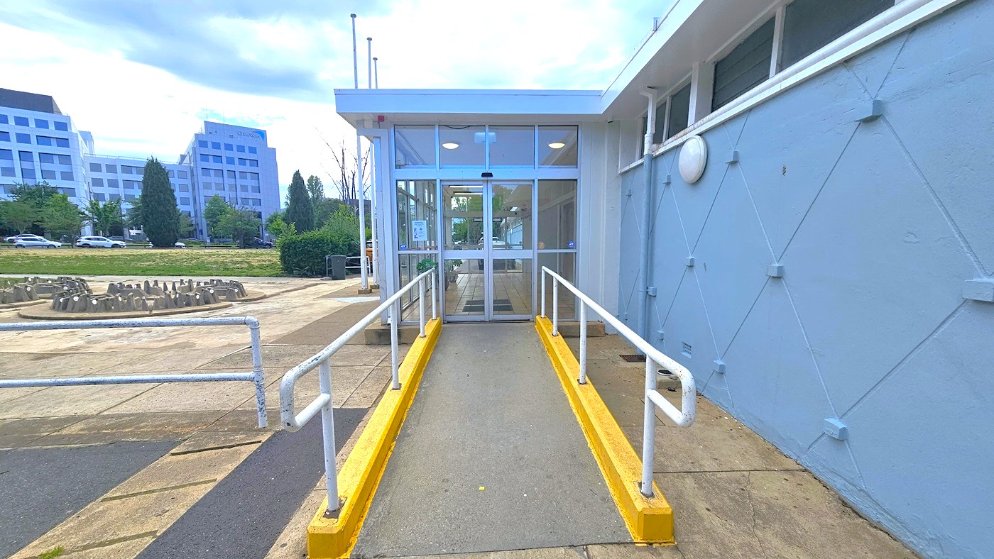 On the left, a concrete bike rack is positioned. A ramp with handrails providing access to the entrance is in the middle. To the right, a blue concrete wall with a diamond-shaped design is present. The building entrance features automatic glass doors. On the far left, there are trees, tall commercial buildings, and green grass visible. On the left, a concrete bike rack is positioned. A ramp with handrails providing access to the entrance is in the middle. To the right, a blue concrete wall with a diamond-shaped design is present. The building entrance features automatic glass doors. On the far left, there are trees, tall commercial buildings, and green grass visible.