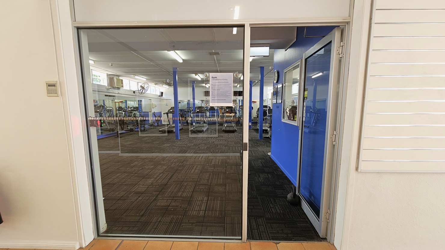 A manual glass door that opens inward leads to the gym. A poster bearing a Gym use reminder is posted on the glass wall. The external wall is all white with a linear design. Inside, the floor is covered with grey carpet. To the right is an office with a glass window and blue walls. A wall clock and framed posters are affixed to the wall. Various gym equipment is visible inside. A manual glass door that opens inward leads to the gym. A poster bearing a Gym use reminder is posted on the glass wall. The external wall is all white with a linear design. Inside, the floor is covered with grey carpet. To the right is an office with a glass window and blue walls. A wall clock and framed posters are affixed to the wall. Various gym equipment is visible inside.