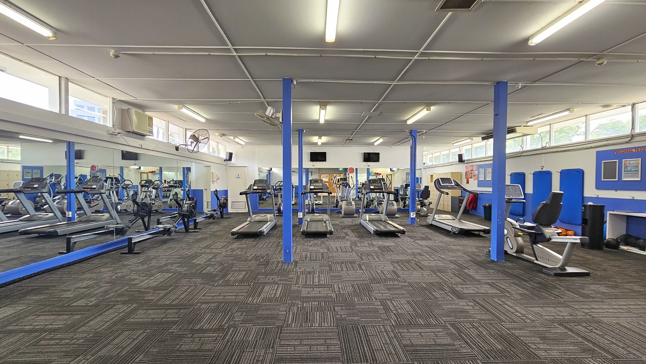 In the middle are various pieces of equipment, including treadmills and rowing machines. The left wall features floor-to-ceiling glass. To the right is a blue and white wall featuring a board with posters. The upper wall features glass windows allowing natural light and a view outside. Flat-screen TVs are mounted on the walls, along with wall fans. The ceiling is all white with fluorescent lights, and the floor has grey tiles. In the middle are various pieces of equipment, including treadmills and rowing machines. The left wall features floor-to-ceiling glass. To the right is a blue and white wall featuring a board with posters. The upper wall features glass windows allowing natural light and a view outside. Flat-screen TVs are mounted on the walls, along with wall fans. The ceiling is all white with fluorescent lights, and the floor has grey tiles.