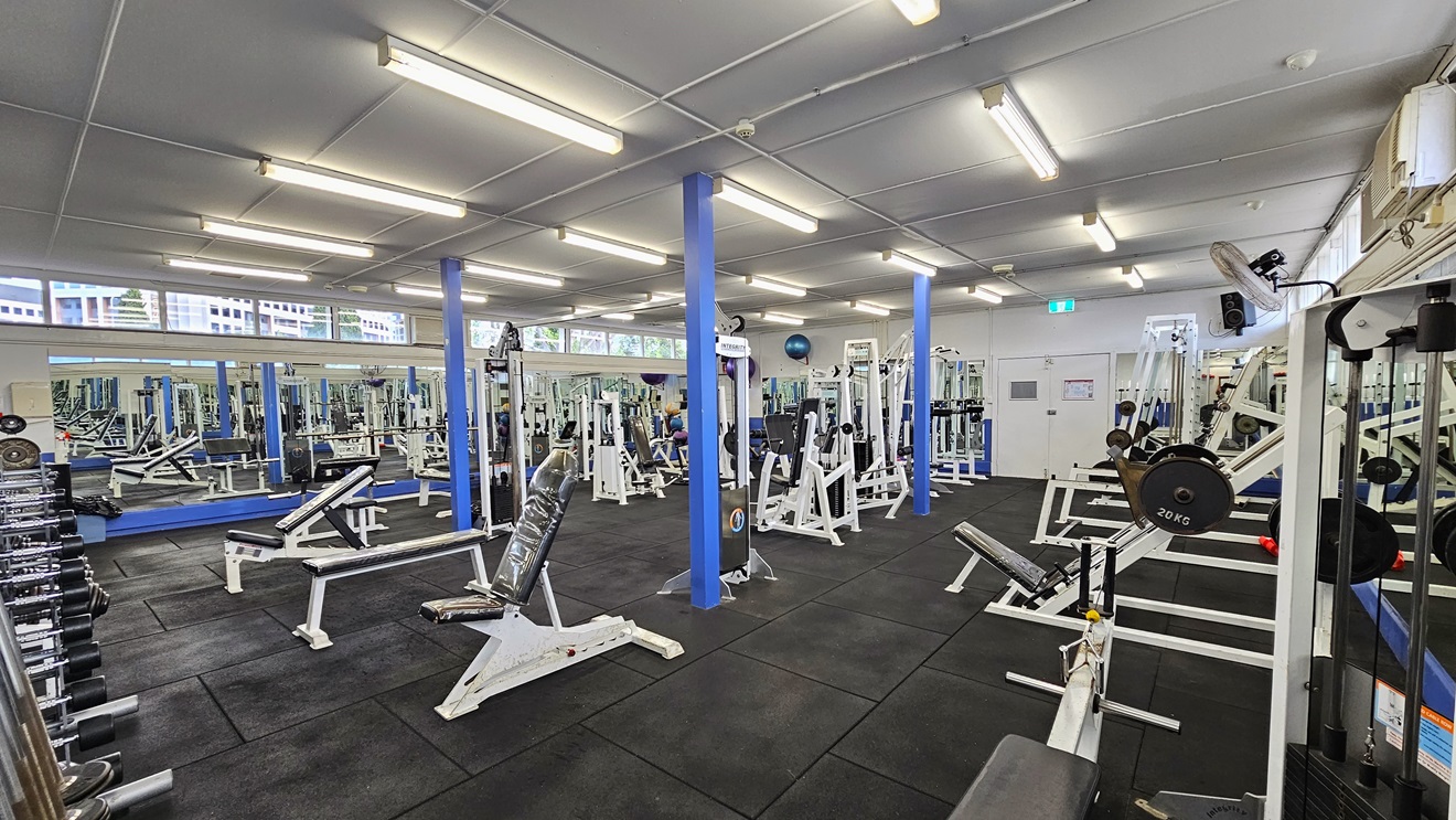 Weights are arranged on the left, and a floor-to-ceiling mirror covers the left wall. Bench chairs, Bench Press, and Smith machines are arranged in the middle and on the right side. The upper wall features glass windows allowing natural light and a view outside. Flat-screen TVs are mounted on the walls, along with wall fans. The ceiling is all white with fluorescent lights, and the floor has black rubber tiles. Weights are arranged on the left, and a floor-to-ceiling mirror covers the left wall. Bench chairs, Bench Press, and Smith machines are arranged in the middle and on the right side. The upper wall features glass windows allowing natural light and a view outside. Flat-screen TVs are mounted on the walls, along with wall fans. The ceiling is all white with fluorescent lights, and the floor has black rubber tiles.