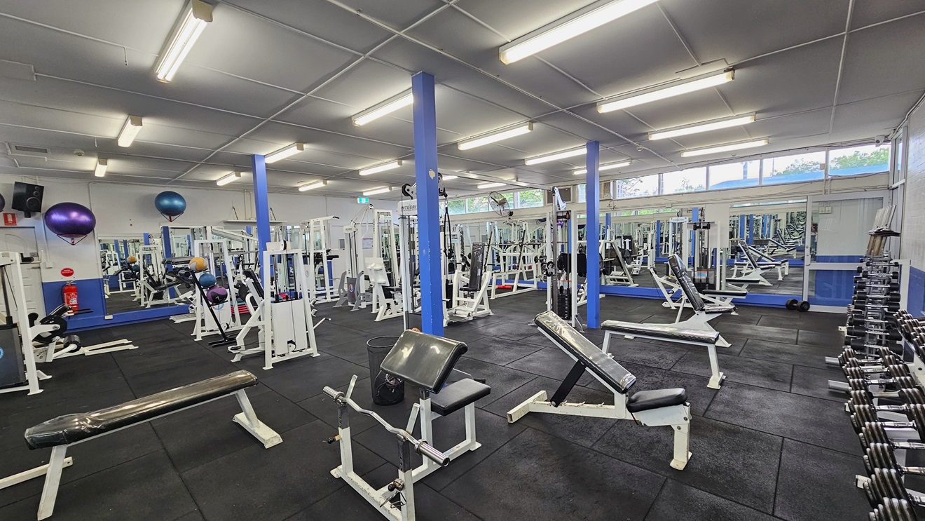 Weights are arranged on the right, and a floor-to-ceiling mirror covers the middle wall. Bench chairs, Bench Press, and Smith machines are arranged in the middle and on the right side. The upper wall features glass windows allowing natural light and a view outside. Flat-screen TVs are mounted on the left wall, along with wall fans. Stability balls are also displayed on the wall. The ceiling is all white with fluorescent lights, and the floor has black rubber tiles. Weights are arranged on the right, and a floor-to-ceiling mirror covers the middle wall. Bench chairs, Bench Press, and Smith machines are arranged in the middle and on the right side. The upper wall features glass windows allowing natural light and a view outside. Flat-screen TVs are mounted on the left wall, along with wall fans. Stability balls are also displayed on the wall. The ceiling is all white with fluorescent lights, and the floor has black rubber tiles.