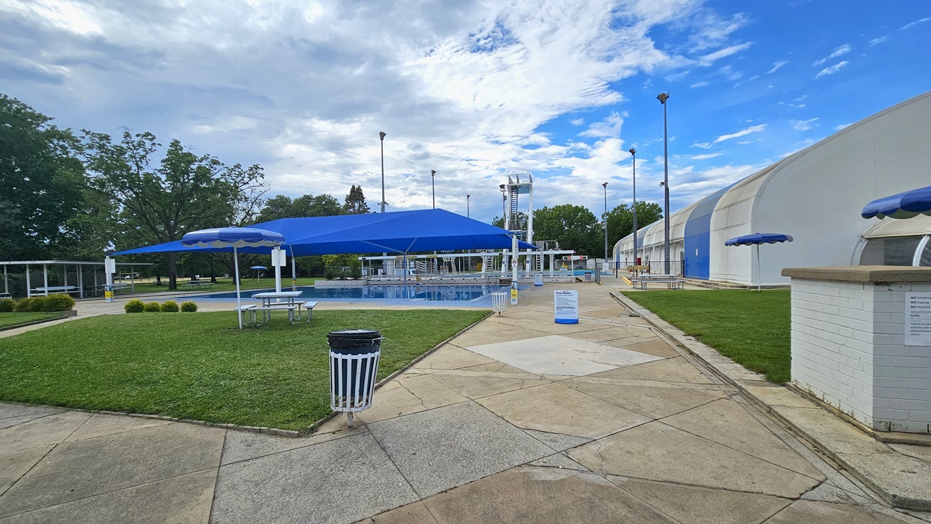 To the left, a pool covered with shade cloth. A picnic table is positioned under an umbrella shade in the manicured grass area. To the right is the dome-shaped indoor aquatic facility building. Trimmed grass also covers the right space. Further ahead, there is a shaded facility with bench chairs, and the diving pool is behind. To the left, a pool covered with shade cloth. A picnic table is positioned under an umbrella shade in the manicured grass area. To the right is the dome-shaped indoor aquatic facility building. Trimmed grass also covers the right space. Further ahead, there is a shaded facility with bench chairs, and the diving pool is behind.