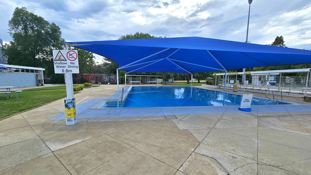 A pool is covered with a blue shade cloth. To the left is a stairs entry equipped with handrails. To the right is another handrail. Safety reminder posters are posted on the post at the left, and a free-standing banner stands in the pool deck. A blue facility building, along with a picnic table, is positioned on the left. To the far end is a shaded tiered seating. The floor is made of concrete. Trimmed grass and tall trees surround the area. A pool is covered with a blue shade cloth. To the left is a stairs entry equipped with handrails. To the right is another handrail. Safety reminder posters are posted on the post at the left, and a free-standing banner stands in the pool deck. A blue facility building, along with a picnic table, is positioned on the left. To the far end is a shaded tiered seating. The floor is made of concrete. Trimmed grass and tall trees surround the area.