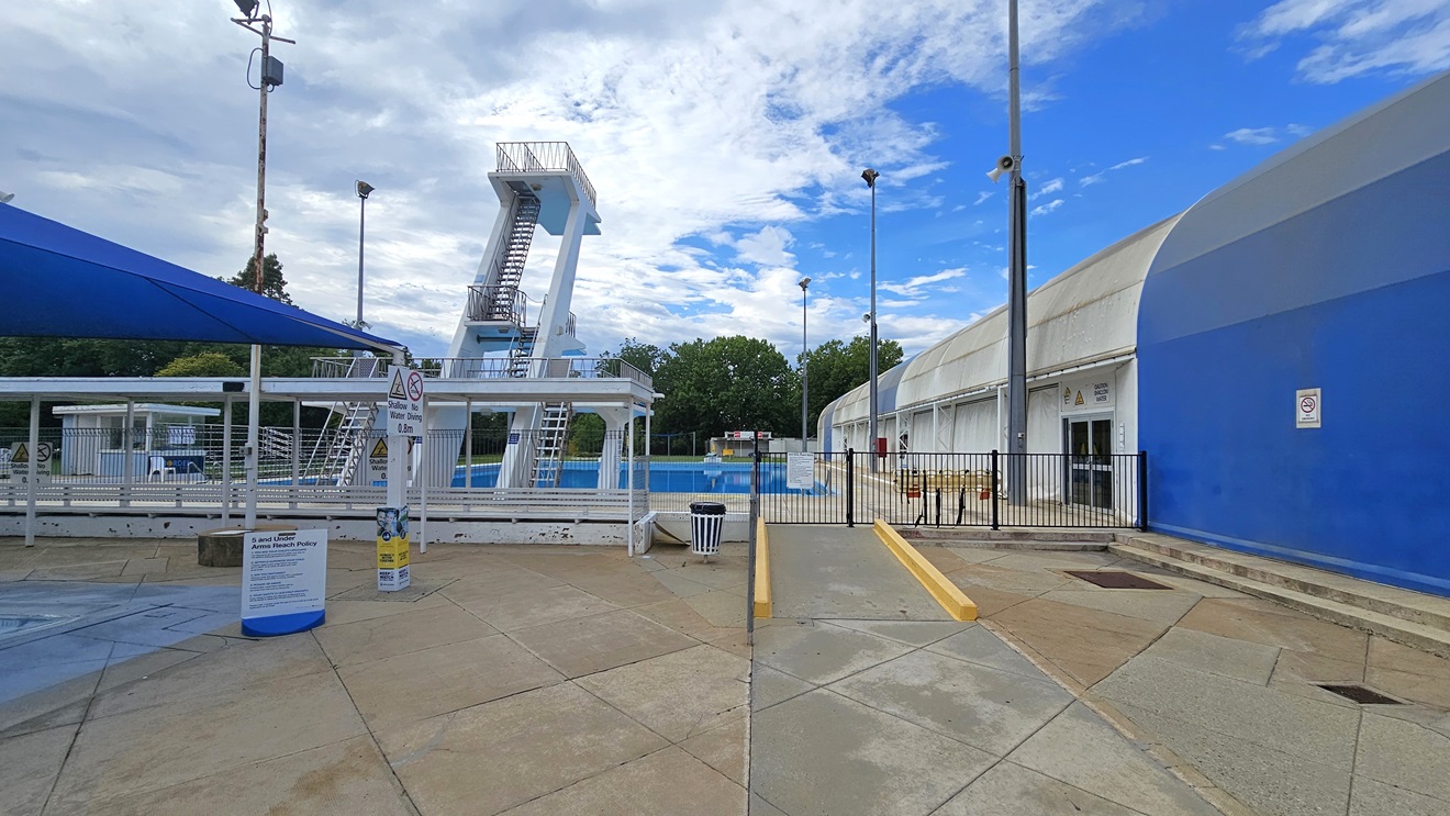 To the left is a section of the kids' pool, and to the right is a ramp leading to the diving pool. On the left is the dome-shaped blue and white structure housing the indoor aquatic center. The diving pool features four-level diving platforms. A steel gate and fence separate the diving pool from the kids' pool. A rubbish bin is positioned near the ramp. Safety banners are affixed to the wall. On the far left is a shaded facility with tiered seating. Tall outdoor lights are visible. To the left is a section of the kids' pool, and to the right is a ramp leading to the diving pool. On the left is the dome-shaped blue and white structure housing the indoor aquatic center. The diving pool features four-level diving platforms. A steel gate and fence separate the diving pool from the kids' pool. A rubbish bin is positioned near the ramp. Safety banners are affixed to the wall. On the far left is a shaded facility with tiered seating. Tall outdoor lights are visible.