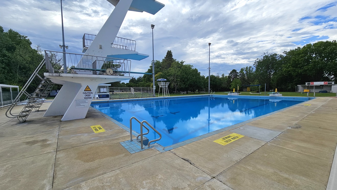 The diving pool has four-level diving platforms with steel stairs and railings. A steel gate and fence separate the diving pool from the kids' pool. The pool is equipped with stairs and handrails, and a security warning denotes the water's depth. To the left is a shaded facility with tiered seating, and adjacent to it is a watchtower. To the far right is a spacious area with green grass and tall trees. Tall outdoor lights are visible. The diving pool has four-level diving platforms with steel stairs and railings. A steel gate and fence separate the diving pool from the kids' pool. The pool is equipped with stairs and handrails, and a security warning denotes the water's depth. To the left is a shaded facility with tiered seating, and adjacent to it is a watchtower. To the far right is a spacious area with green grass and tall trees. Tall outdoor lights are visible.