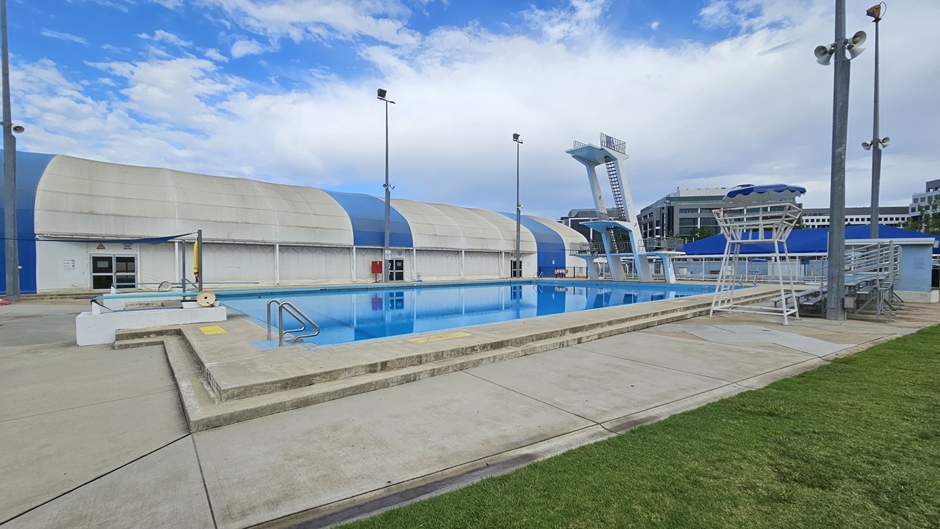 The pool is equipped with stairs and handrails. To the right is a watchtower, and next to it is tiered seating. To the left is the blue-white dome-shaped building housing the indoor aquatic facilities. On the left is a shaded facility with tiered seating, and adjacent to it is a watchtower. The diving platform is located in the middle, equipped with steel stairs and railings. Neatly trimmed grass surrounds the right side of the pool. The pool is equipped with stairs and handrails. To the right is a watchtower, and next to it is tiered seating. To the left is the blue-white dome-shaped building housing the indoor aquatic facilities. On the left is a shaded facility with tiered seating, and adjacent to it is a watchtower. The diving platform is located in the middle, equipped with steel stairs and railings. Neatly trimmed grass surrounds the right side of the pool.