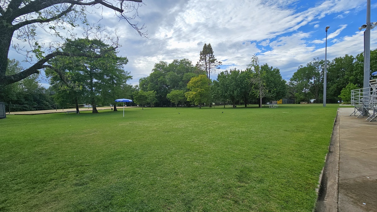 To the left is a spacious area covered with green grass and tall trees. Picnic tables are placed under the trees. An umbrella shade is visible. To the right is tiered seating and the watchtower. To the left is a spacious area covered with green grass and tall trees. Picnic tables are placed under the trees. An umbrella shade is visible. To the right is tiered seating and the watchtower.