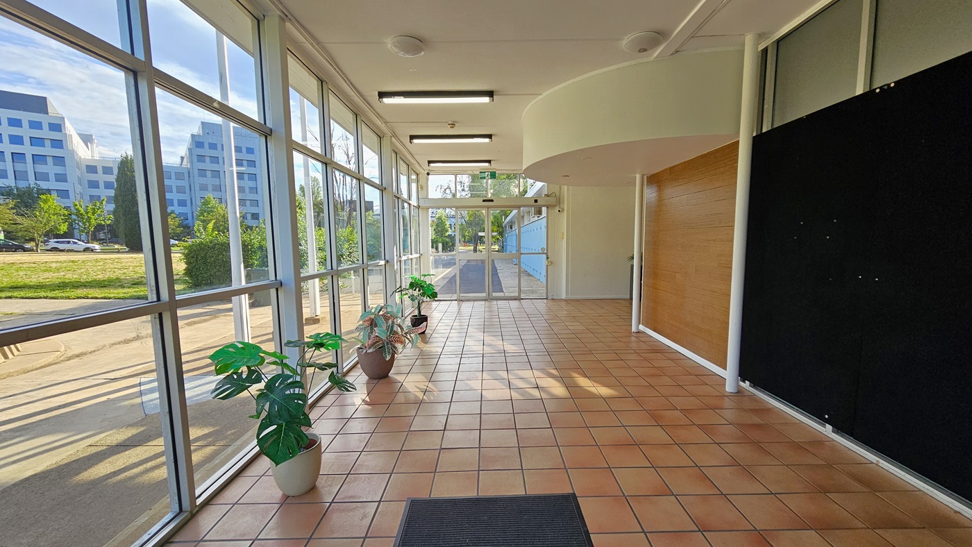 The doorway features a black anti-slip mat. The left side is glass, offering an exterior view. Potted ornamental plants line up along the wall. The right wall combines brown concrete and brown tiles. At the corridor's end is another glass door. The ceiling is uniformly white with fluorescent and LED lighting, and the floor is covered in brown tiles. The doorway features a black anti-slip mat. The left side is glass, offering an exterior view. Potted ornamental plants line up along the wall. The right wall combines brown concrete and brown tiles. At the corridor's end is another glass door. The ceiling is uniformly white with fluorescent and LED lighting, and the floor is covered in brown tiles.